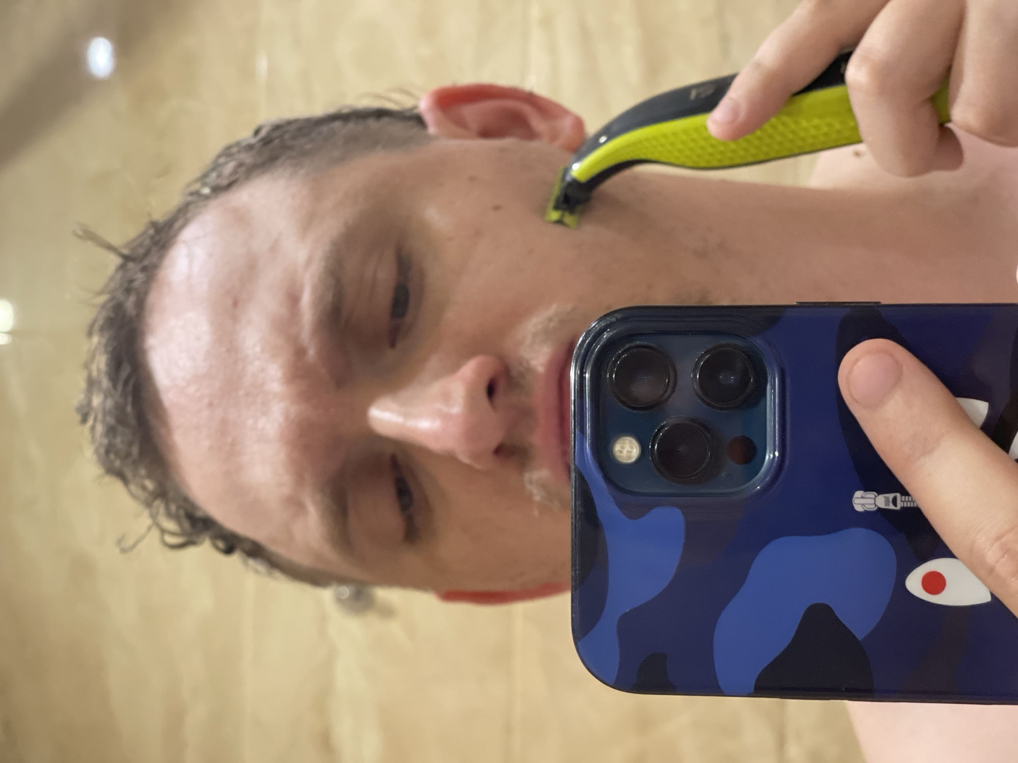 In this close-up selfie, a person is seen shaving their face with an electric razor. The background appears to be a bathroom with tiled walls. The person is holding a smartphone with a blue camouflage case to capture the image. The lighting suggests an indoor environment. The location of Khimki, Russia is not depicted in the image.