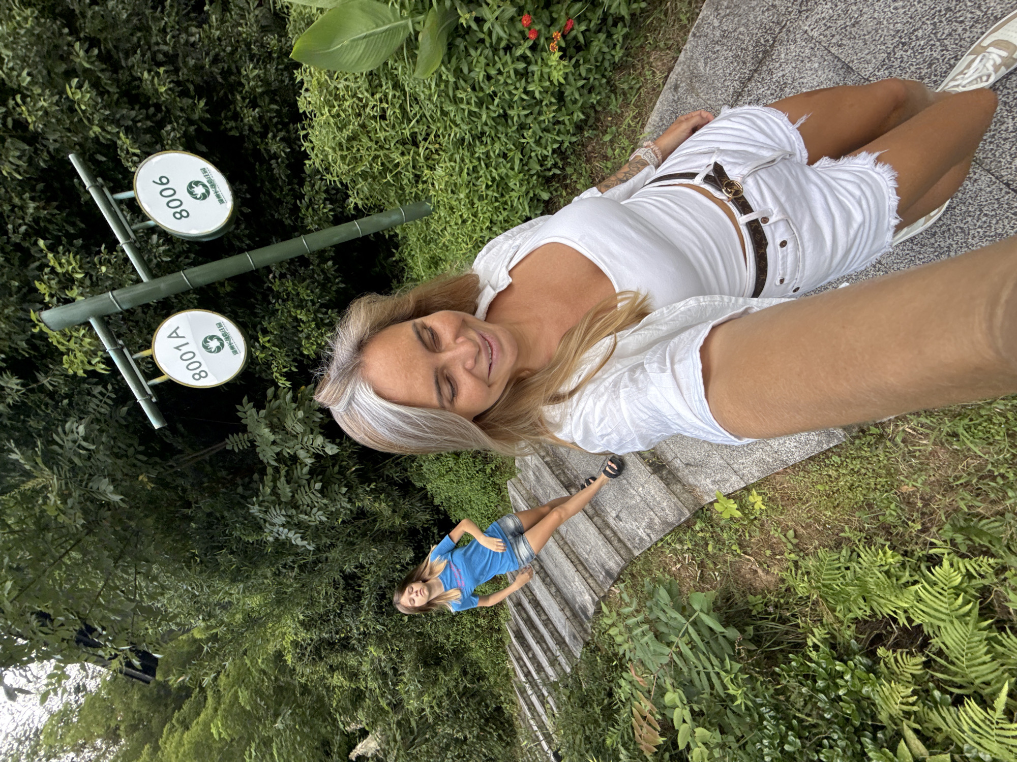 In Wuxing District, China, two individuals are captured on a paved pathway ascending a stone staircase surrounded by lush green foliage. The foreground features a woman with blonde hair, wearing a white top and white shorts, holding a phone to take a selfie. Behind her, a younger woman with blonde hair is positioned further up the stairs, wearing a blue t-shirt and denim shorts. Two circular signs are visible on a bamboo pole, one labeled "8001A" and the other "8006," both featuring a green logo. The overall environment is natural and verdant, suggesting a park or outdoor recreational area.