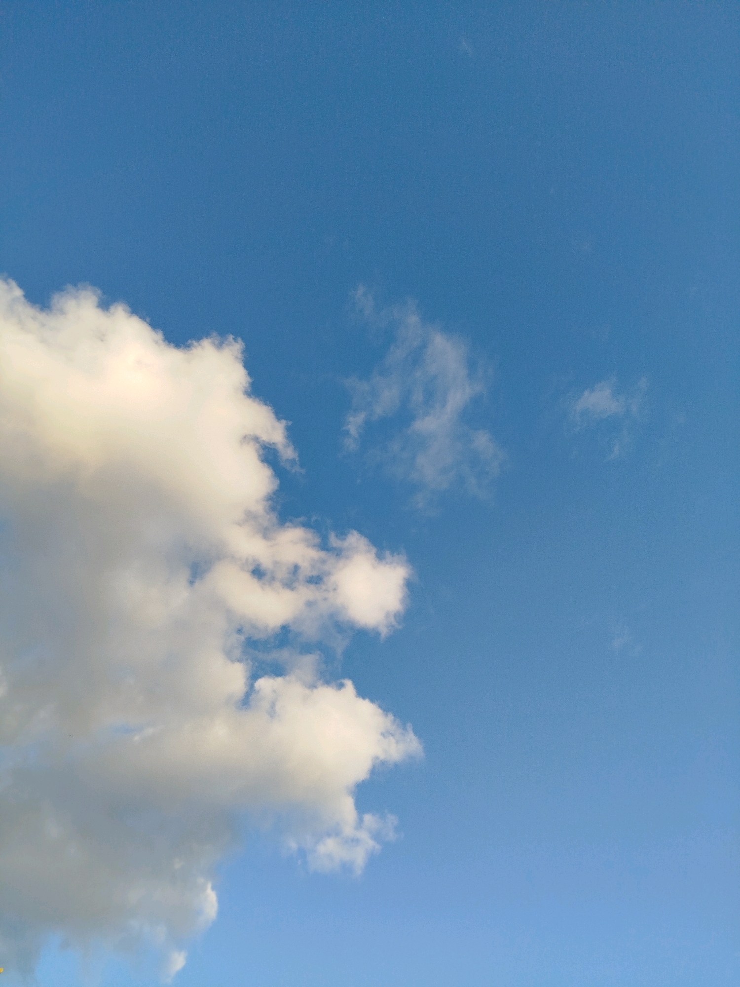 This is a close-up shot of a bright blue sky with scattered white clouds. A large, fluffy cloud dominates the left side of the frame, with smaller, wispy clouds visible in the distance. The lighting suggests it is daytime, and the clear sky indicates fair weather. There are no buildings, people, or any other objects in the frame to provide context about a specific location within Mirny, Russia.