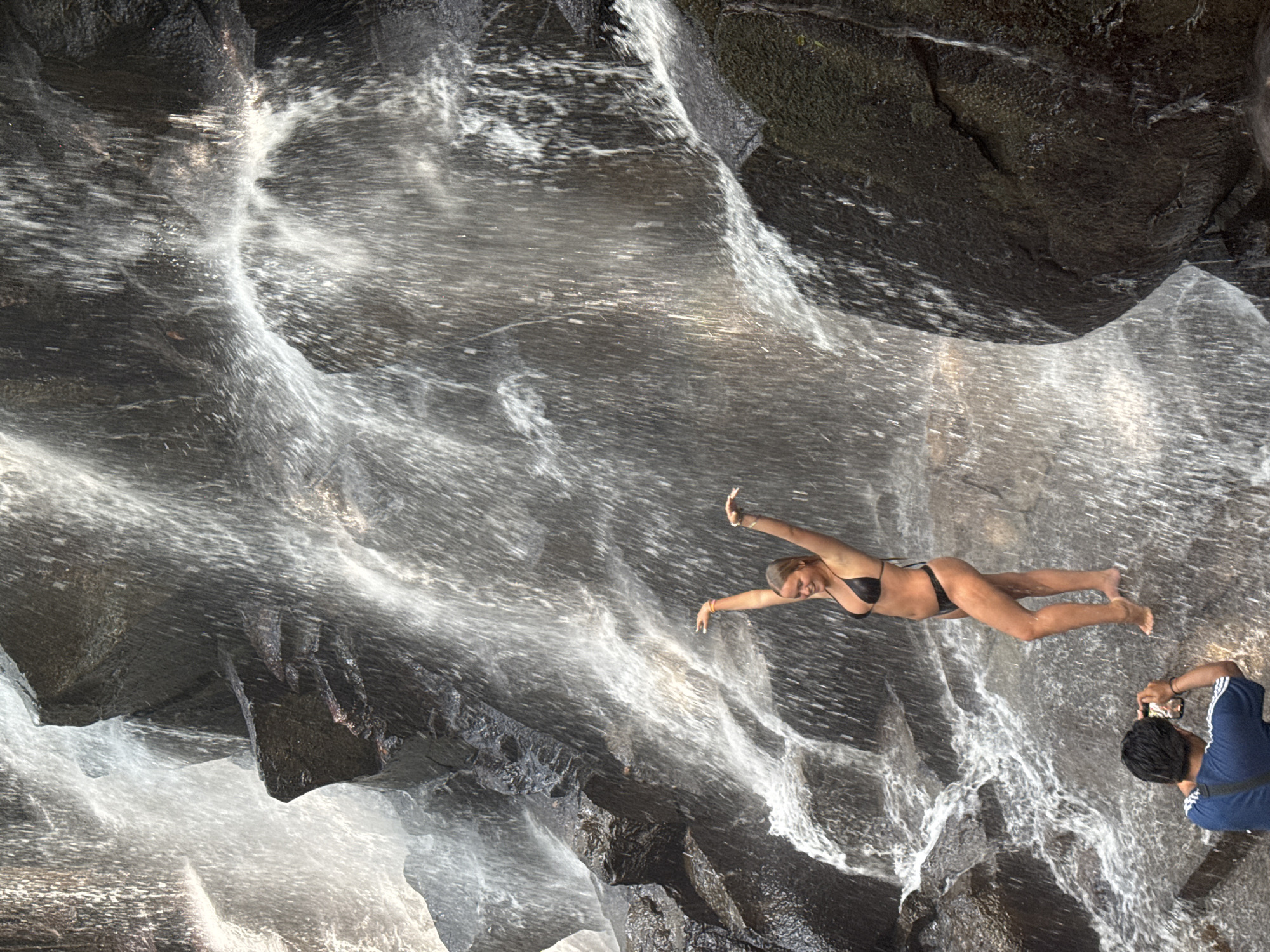 A young woman in a black bikini poses with her arms outstretched under a cascading waterfall. She stands on wet rocks, her body angled slightly towards the viewer. To her left and lower, a person in a blue t-shirt holds up a phone to take a picture of her. The waterfall flows over dark, mossy rocks, creating a misty and dynamic environment. The location is a natural setting with no identifiable urban features.
