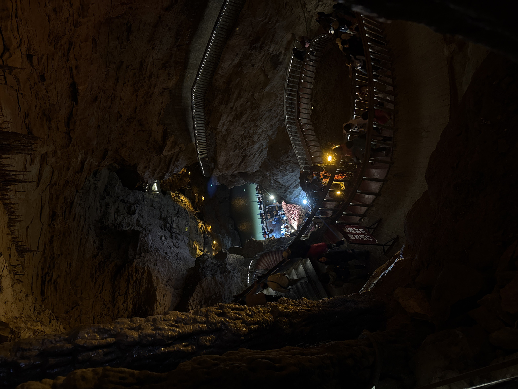 The image shows a dark cave with a metal walkway winding through it. People are walking on the walkway. There is a body of water at the end of the path. The location is in the city of Unknown, Unknown.