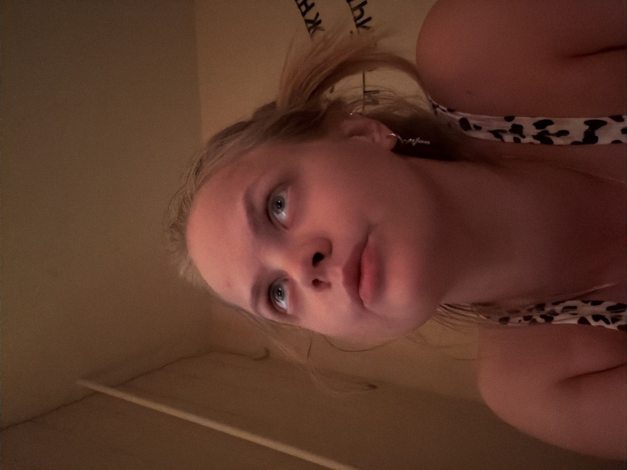 A selfie photograph depicts a young, fair-skinned woman with blonde hair partially pulled back. She has blue eyes and is looking upwards. She is wearing an earring and a top with a black and white pattern. The background consists of light brown walls and ceiling with a white molding. The exact location within the city of Izhevsk, Russia is not discernible from this image.