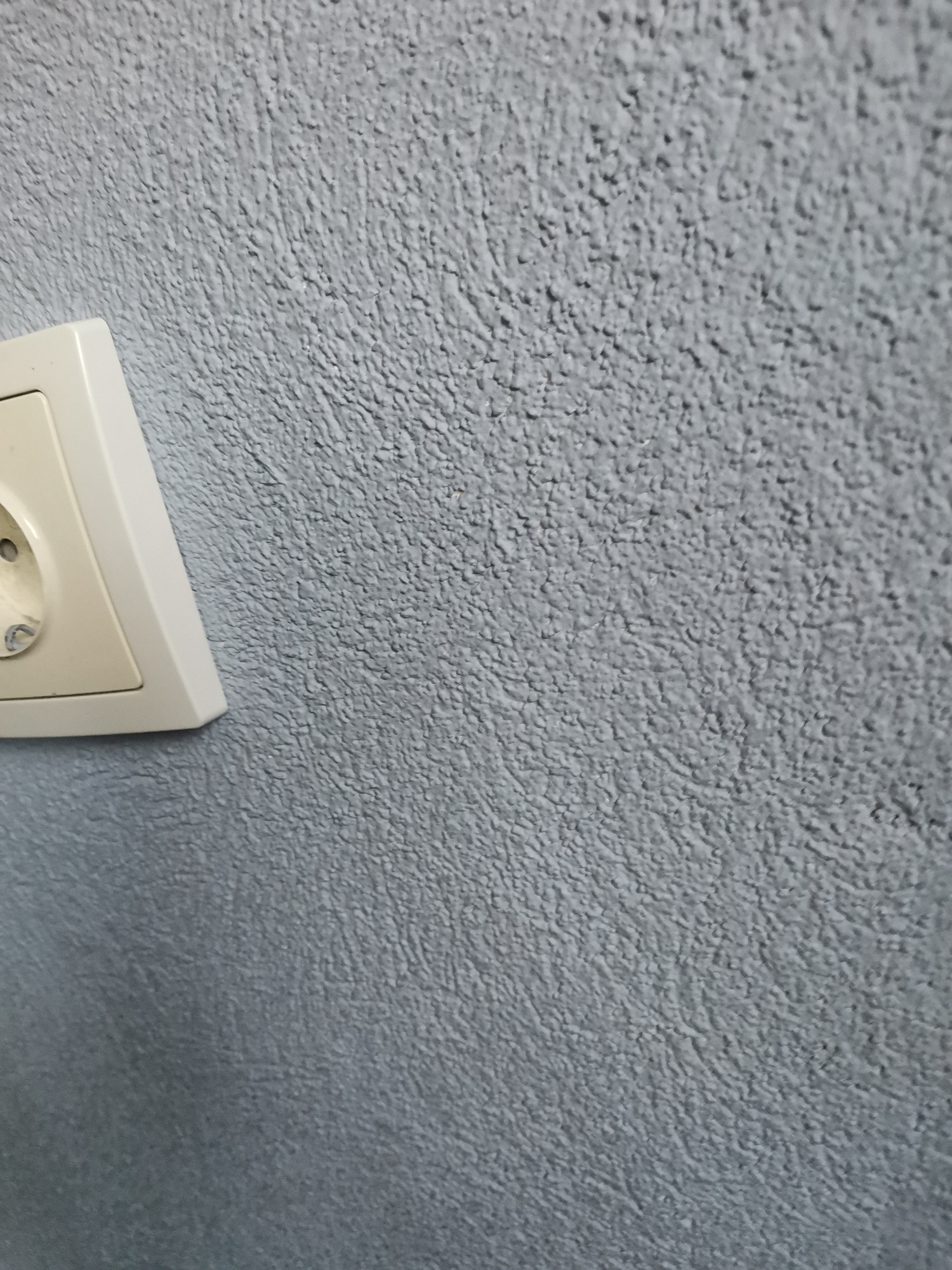 This image presents an interior wall within a structure located in Pokrovnik, Bulgaria. The wall is coated in a light blue-grey textured finish, displaying an irregular, granular surface. Partially visible on the lower-left is an off-white Type F electrical socket, installed flush with the wall. The visible segment includes one circular pin receptacle and a portion of the integrated grounding clip. No individuals or dynamic actions are present. Illumination appears ambient and uniform.