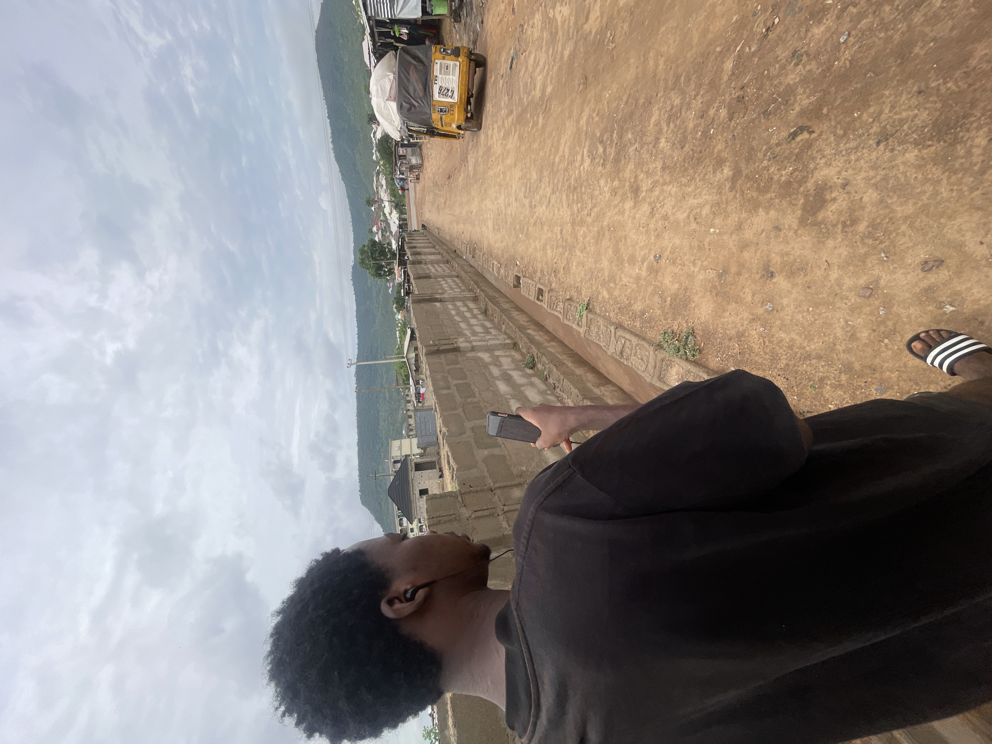 A low-angle shot looking down a street in Lokoja, Nigeria. The viewpoint is from a slight elevation, possibly a rooftop or raised platform. A man, seen from the chest down, is holding a smartphone in his right hand; he is wearing a black T-shirt and Adidas sandals. The street is unpaved, showing exposed dirt. A yellow, three-wheeled vehicle (a "keke") is visible further down the road. Buildings and lush green hills appear in the background. Overcast skies are visible.