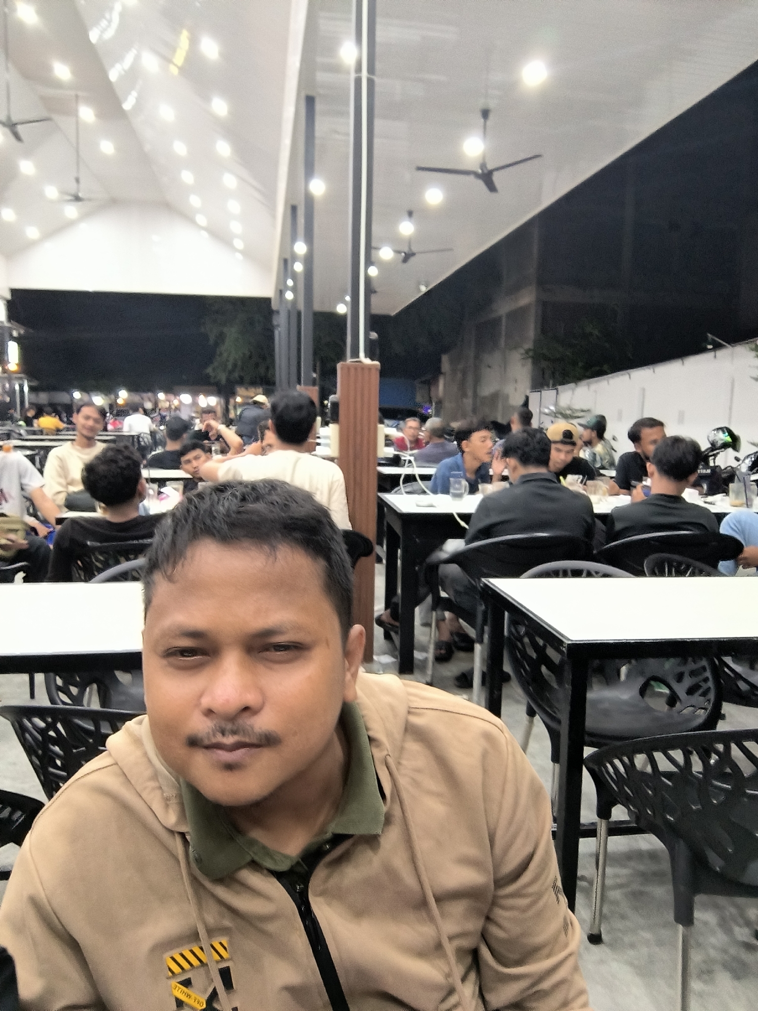 A close-up, eye-level shot shows a man in the foreground looking towards the camera. He has dark hair, a mustache, and is wearing a tan hooded jacket over a green shirt. Behind him, several other individuals are seated at tables in an outdoor dining area. The environment appears to be a covered space with a white ceiling, illuminated by numerous lights. In the background, there are more tables, chairs, and people dining. The overall setting suggests a casual restaurant or food court in Lhokseumawe, Indonesia, possibly at night or in a dimly lit area, given the artificial lighting.