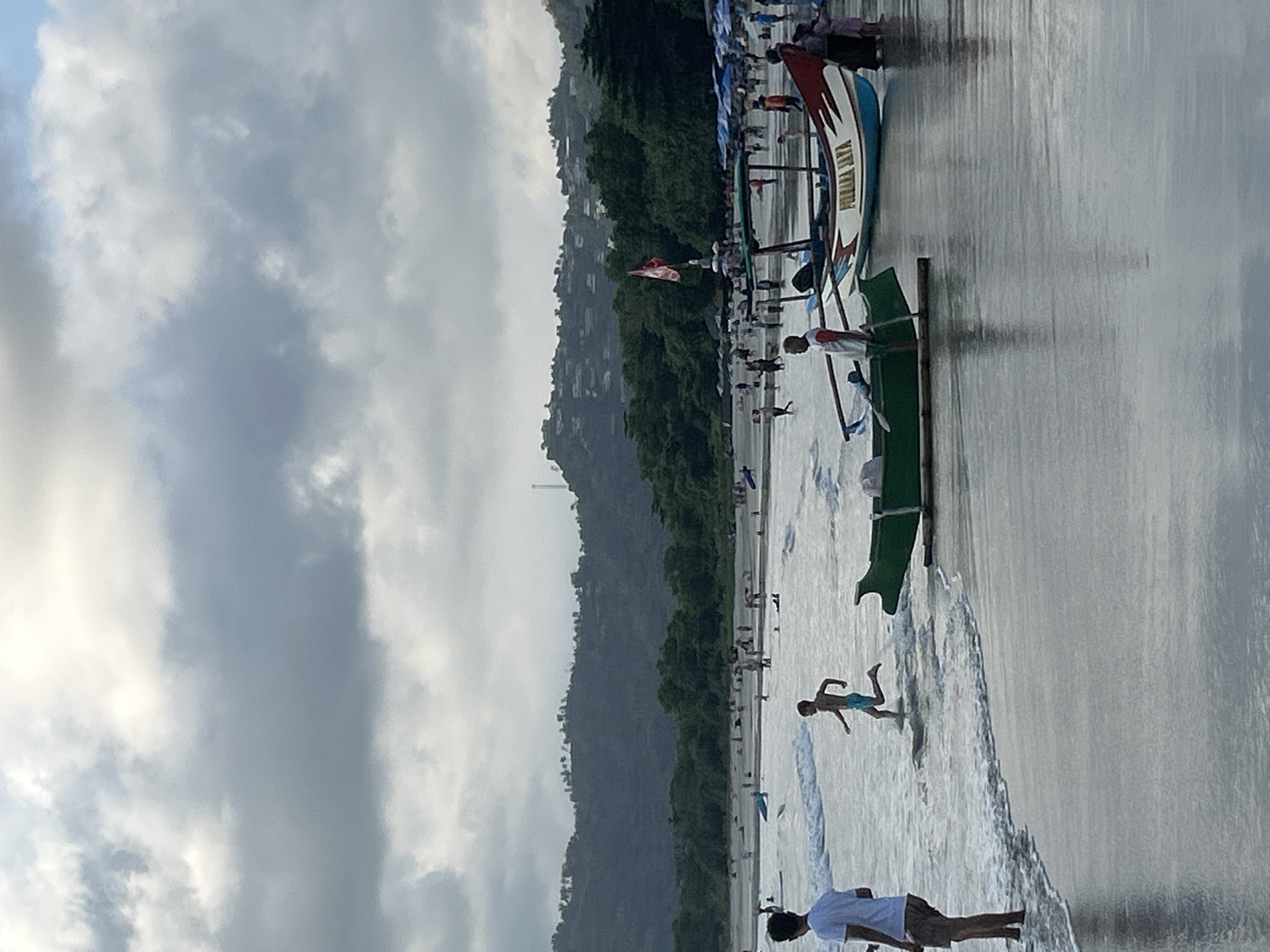 The image depicts Selongbelanak Beach in Selongbelanak, Indonesia, under a cloudy sky. The shoreline is populated with numerous people, some wading in the shallow water and others walking on the wet sand. Several boats are visible, including two traditional outrigger boats in the foreground, one green and one with red and white accents. A red and white flag flies from a pole behind the boats. In the background, lush green hills covered with vegetation and scattered buildings rise, indicating a coastal village setting. The sea appears calm with small waves rolling onto the beach. The overall atmosphere is one of a busy beach day.