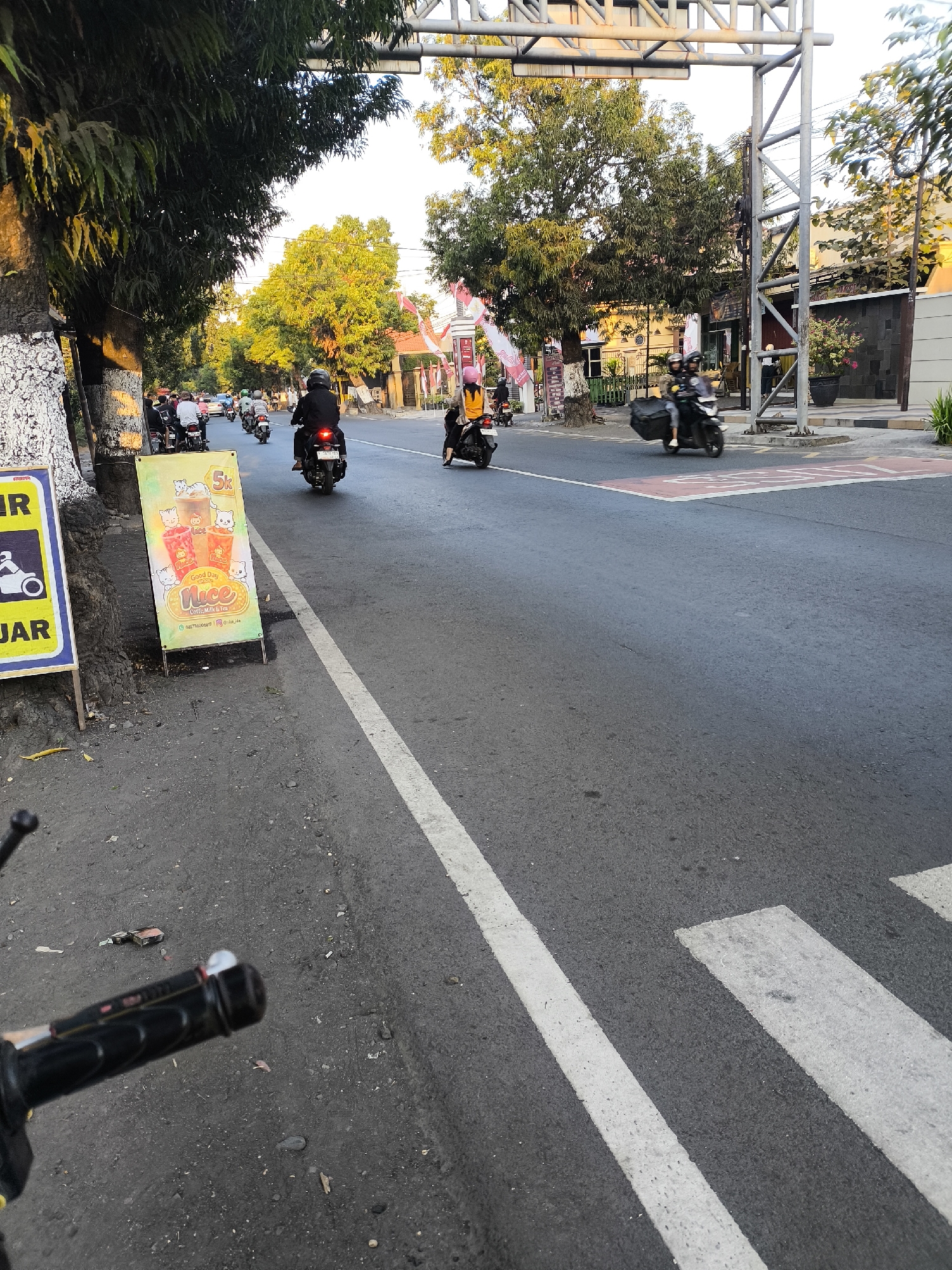 The image shows a street scene in Citrodiwangsan, Indonesia. A road with a white lane divider runs diagonally across the frame. Several people are riding motorcycles, some individually and some with passengers. In the foreground, on the left side of the road, there is a sign advertising "Good Day Nice Coffee Milk & Tea". Trees line the left side of the street, and buildings are visible in the background. The sky is clear and bright.