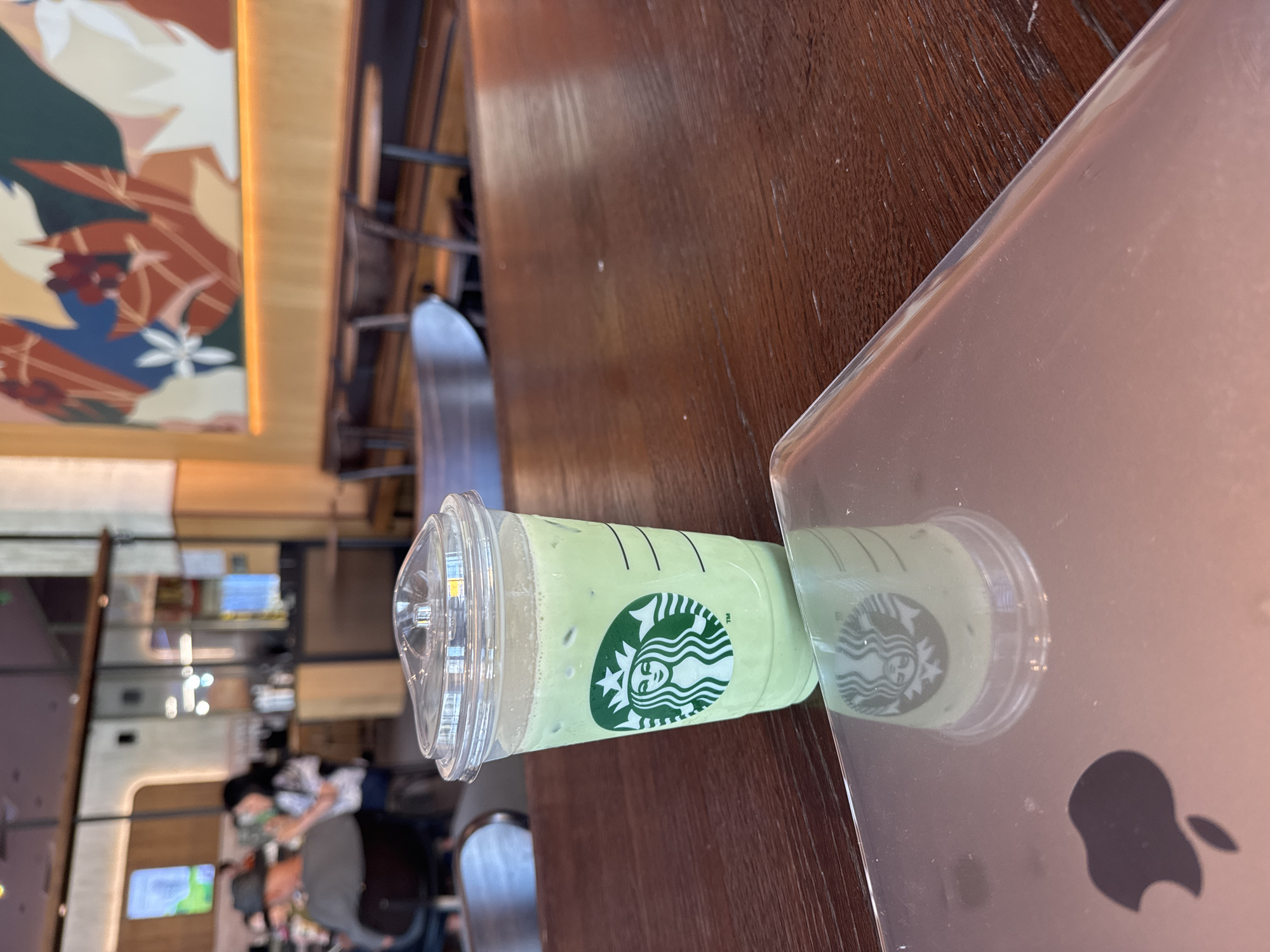 The image depicts a Starbucks iced matcha latte resting on top of an Apple MacBook laptop, which is placed on a wooden table inside a Starbucks coffee shop. The environment is an interior setting with other patrons and architectural details in the background. The city and country, Sofia, Bulgaria are implied because of the coffee shop.