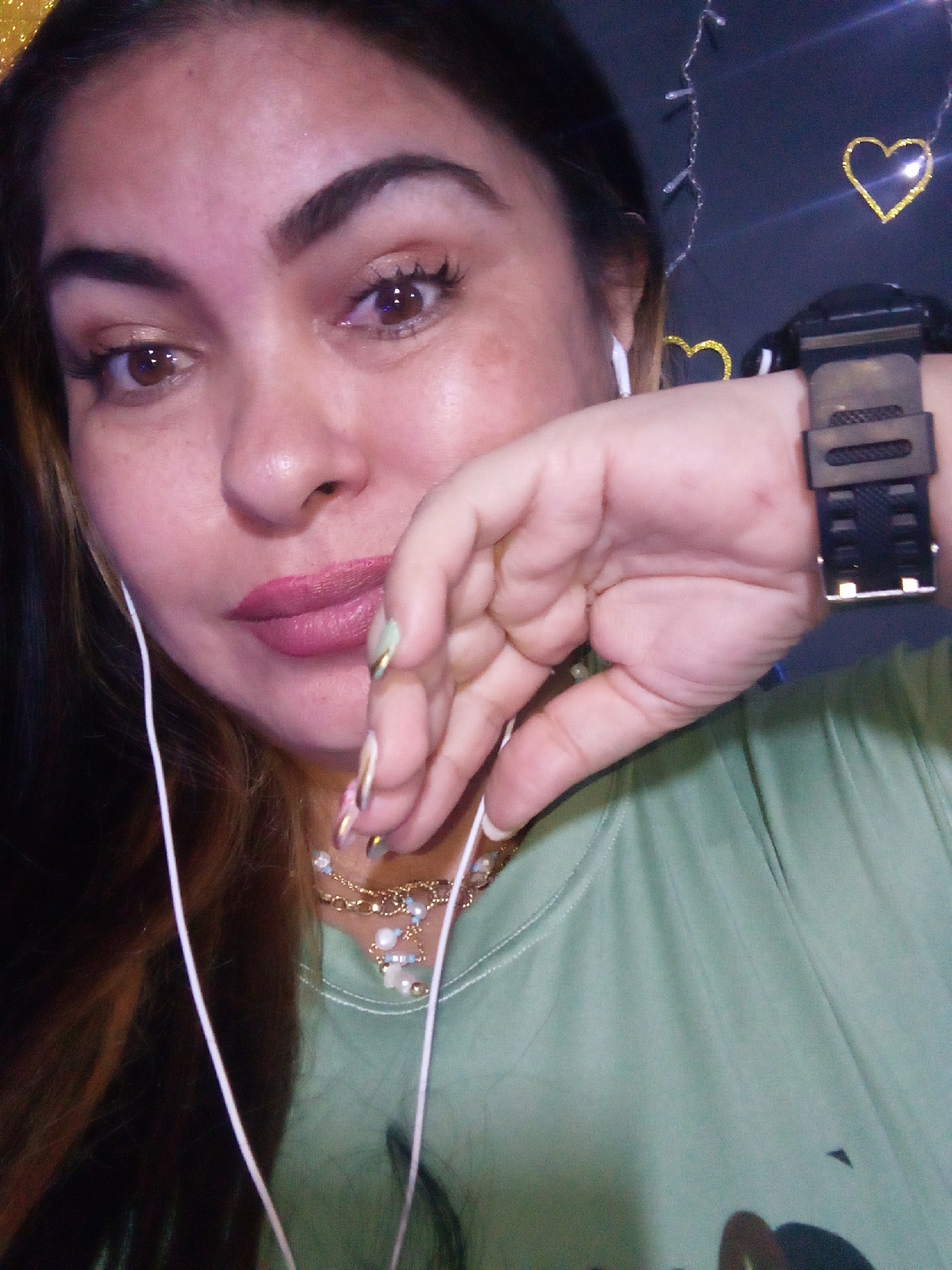 The image displays an adult female from the chest up, positioned with her head slightly tilted to the viewer's left, looking directly at the camera. She has long, dark hair with brown highlights visible on the left side of the frame. Her brown eyes feature visible eye makeup, including eyeshadow, liner, and mascara. She has well-defined dark eyebrows, a prominent nose, and full lips colored with pink/mauve lipstick. Her skin tone is light to medium, with several small blemishes or freckles visible on her face.
She is wearing a light green (sage) round-neck shirt. White wired earphones are inserted into both ears, with the wires extending downwards over her chest. Her right hand (viewer's left) is raised, with fingers partially covering the lower portion of her face, near her chin and mouth. Her fingernails are long and manicured, adorned with light and metallic polish accents. On her left wrist (viewer's right), she wears a black digital watch with a textured rubber strap. A layered necklace, consisting of gold-toned chains, small pearls, and light-colored beads, is visible around her neck.
The background is uniformly dark, possibly a wall or drape. It is decorated with string lights, including a vertical strand of small, clear bulbs on the left side and two gold-colored, glitter-textured heart shapes. One heart is prominent on the right side, while another is partially visible on the lower left. Light reflections are noticeable on the heart decorations and the watch. The scene is located in Juan Griego, Venezuela.