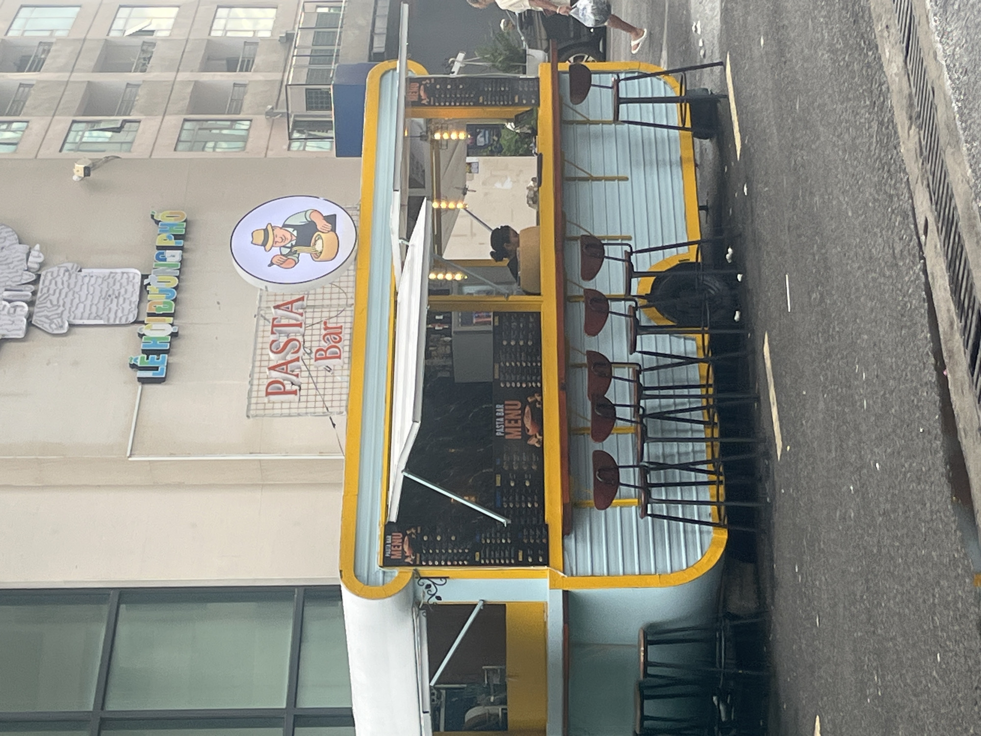 The image shows a "Pasta Bar" stand located in Bac Nha Trang Ward, Vietnam. The stand is a yellow and light blue structure with a menu displayed. Several stools are lined up along the side for customers. The stand is positioned near a building with the name "TE LE DONG PHO" visible. A person is walking in the background. The scene appears to be outdoors on a street.