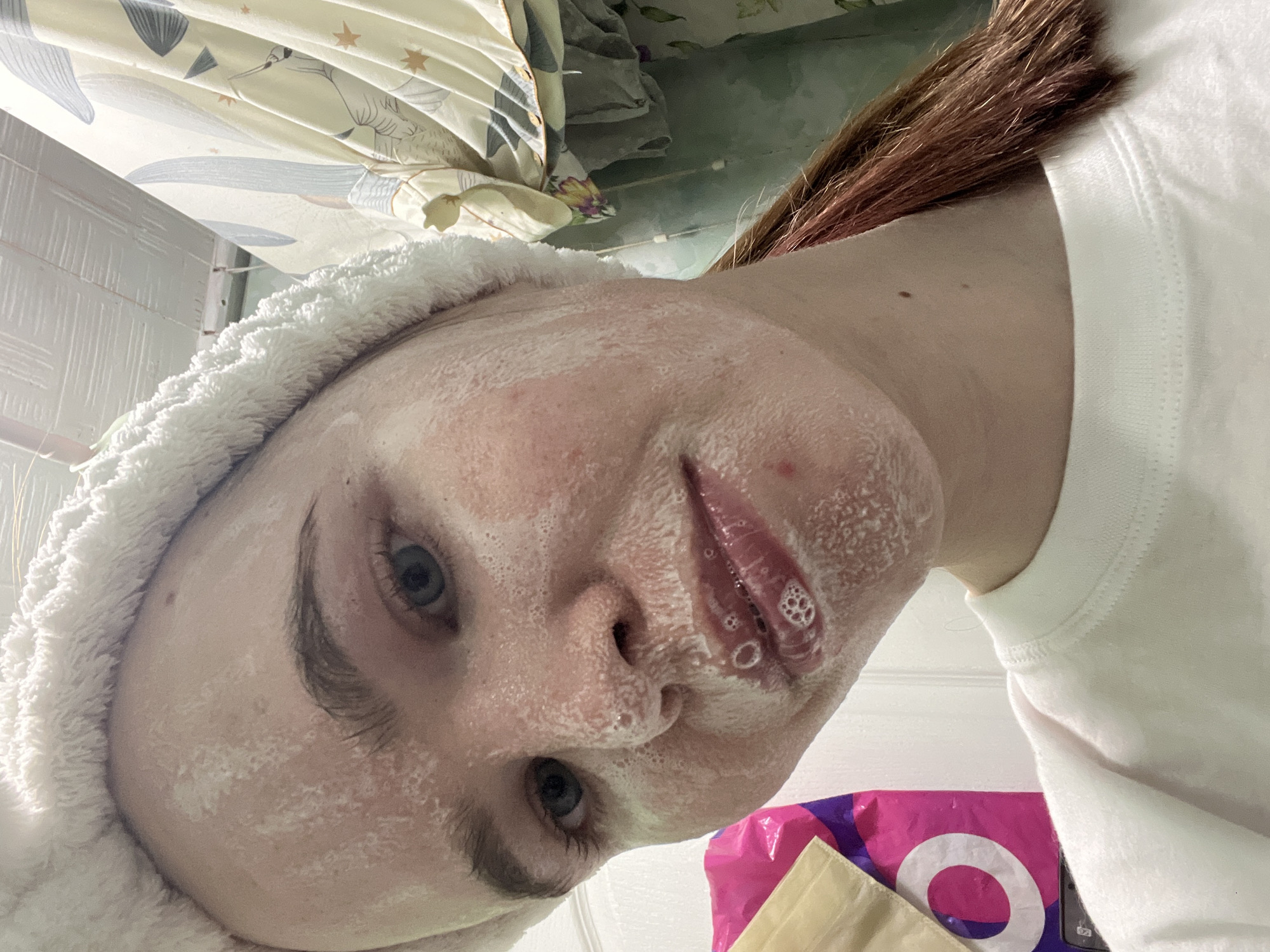 A close-up, tilted selfie shows a woman with a white towel wrapped around her head and a white facial cleanser lathered on her face. The background includes a shower curtain with a bird design, a partially visible wall with white tiles, and a pink shopping bag with a white circle on it. The woman has light skin, brown hair, and light-colored eyes. Bubbles are visible on her lips. The location is likely indoors in a bathroom. The exact location in Chaikovsky, Russia is not discernible from the image.