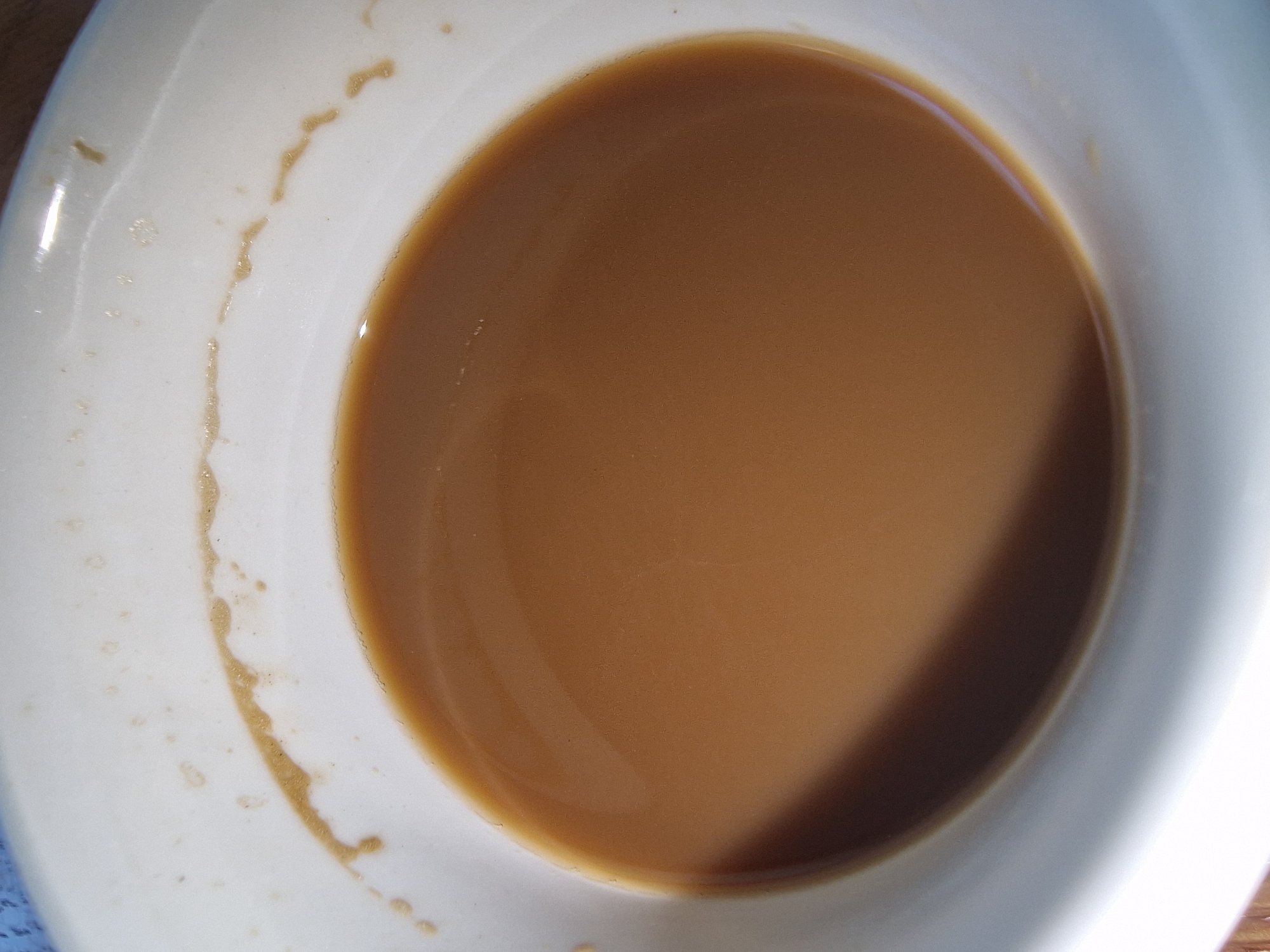 A top-down view of a white ceramic cup partially filled with a light brown liquid, possibly coffee with milk or tea. A dried ring of the same liquid is visible on the inside of the cup near the rim. The location and city are unknown.