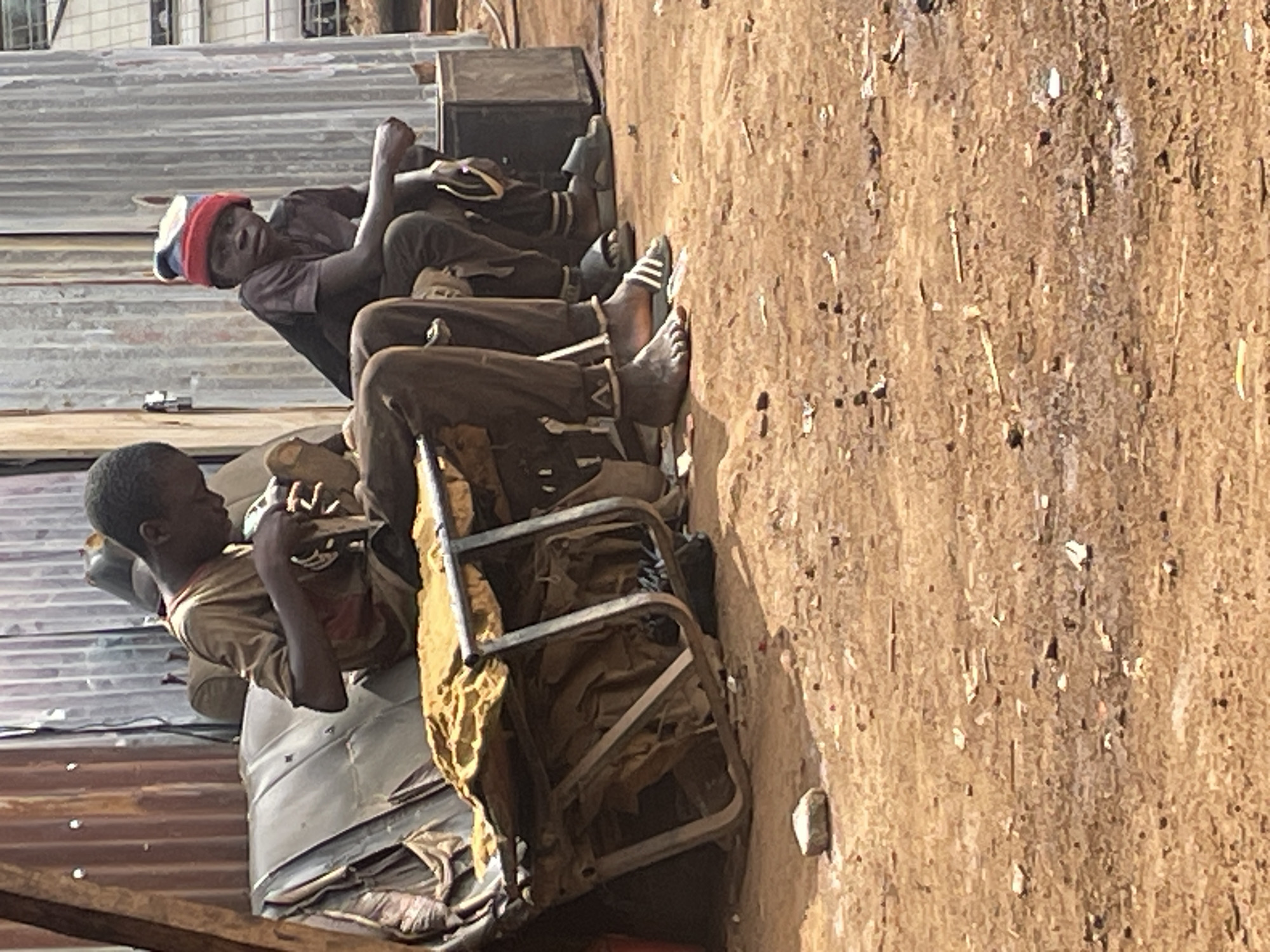 The image shows two young males sitting in makeshift chairs constructed from scrap metal and fabric in Jos, Nigeria. The environment appears to be a poverty-stricken area with corrugated metal roofing and a dirt ground visible. One male wears a red and blue hat, while the other has bare feet. Both are wearing simple clothing. The scene suggests a casual, perhaps resting, moment in an impoverished area.