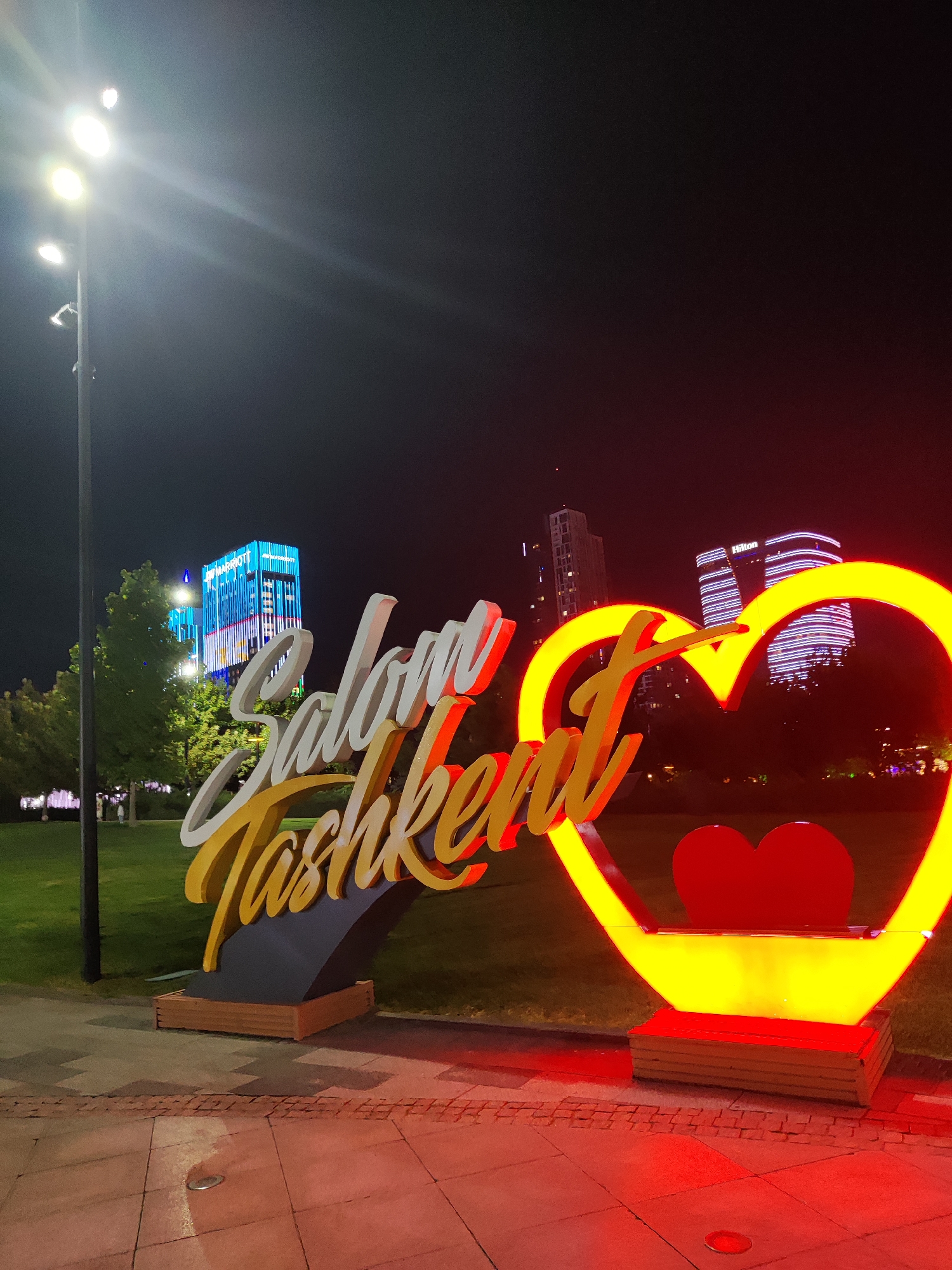 This is a nighttime photograph of a large, illuminated sign in Tashkent, Uzbekistan. The sign features stylized script spelling out "Salom Toshkent" (Hello Tashkent) in white and gold lettering, arching over a bright yellow, illuminated heart shape with a red heart inside. The sign is erected on a dark base with wooden accents. In the background, several modern buildings are visible, including a prominent blue and white illuminated skyscraper with the "JW Marriott" logo, and another building with the "Hilton" logo. The surrounding area appears to be a park or public space, with a manicured green lawn and a paved walkway in the foreground. Streetlights cast bright white light, and the neon illumination of the sign creates a warm red and yellow glow on the ground. No people are visible in the image.