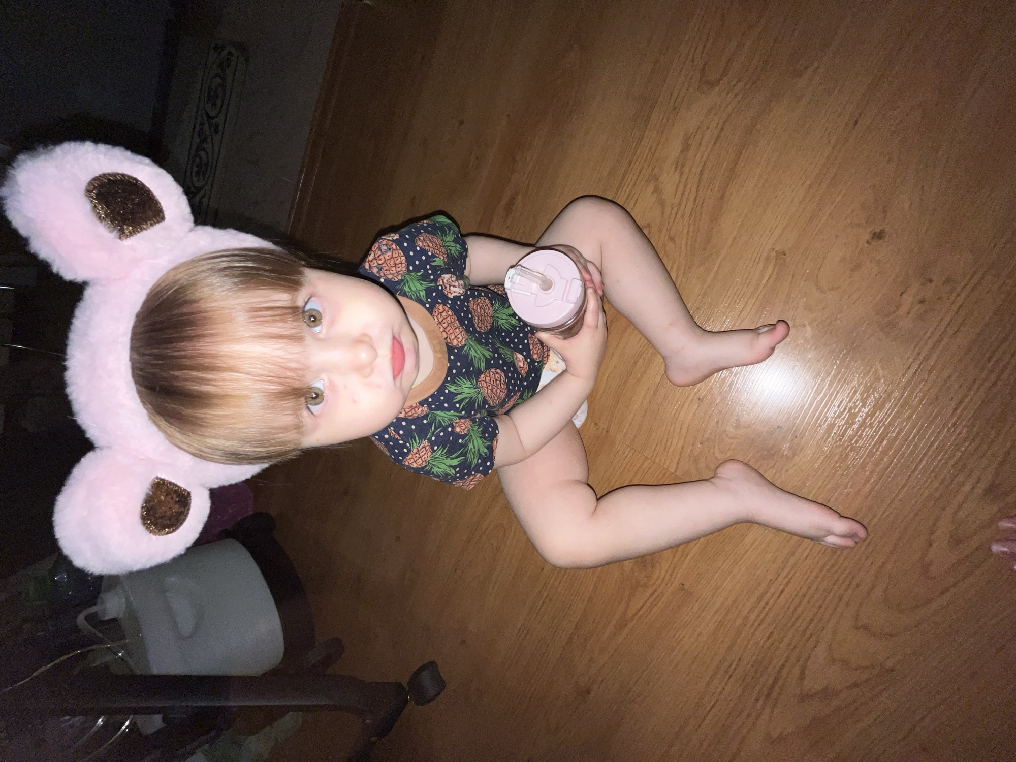 A small child, viewed from above, sits on a wooden floor. The child is wearing a navy blue onesie with a pineapple print and a pink headband with large, fuzzy pink ears that have brown patches. They are holding a pink sippy cup with a straw in their hands. Their legs are outstretched in front of them. A gray container with a black base is visible in the lower left corner. A small portion of a human foot is visible in the lower right corner. The location appears to be indoors.