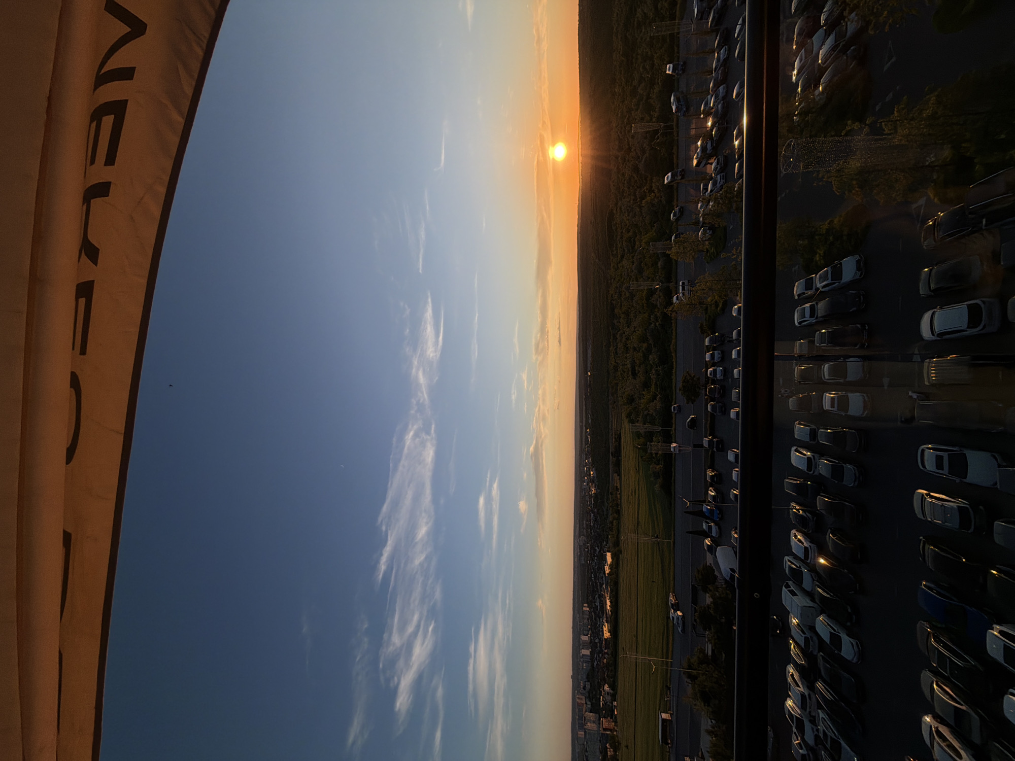 The image captures a panoramic view of Stăuceni, Moldova, during a sunset. The sky is a gradient of blue, with wisps of white clouds and a crescent moon visible in the upper portion. The sun, a bright orange orb, is setting on the horizon, casting a warm glow over the landscape. Below the sky, a city skyline with buildings is visible, transitioning into a vast expanse of green trees. In the foreground, a parking lot is filled with numerous cars, neatly arranged. The scene suggests a peaceful evening in a suburban or semi-urban environment.