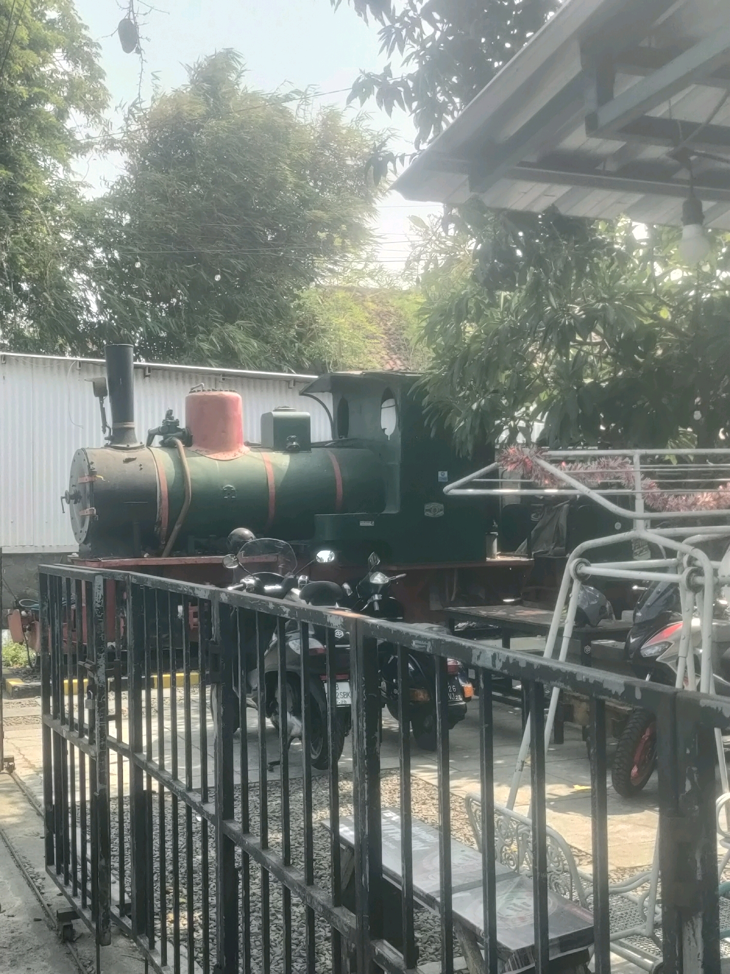 The image depicts a vintage steam locomotive, painted green with a red dome, positioned outdoors in what appears to be a dining or recreational area. A black metal fence in the foreground partially obstructs the view of the locomotive and several motorcycles parked behind it. The background shows lush green trees and a white corrugated metal structure. The scene suggests a unique attraction, possibly in a restaurant or theme park, located in Surabaya, Indonesia, given the context provided. The lighting indicates it is daytime with a slightly overcast sky.