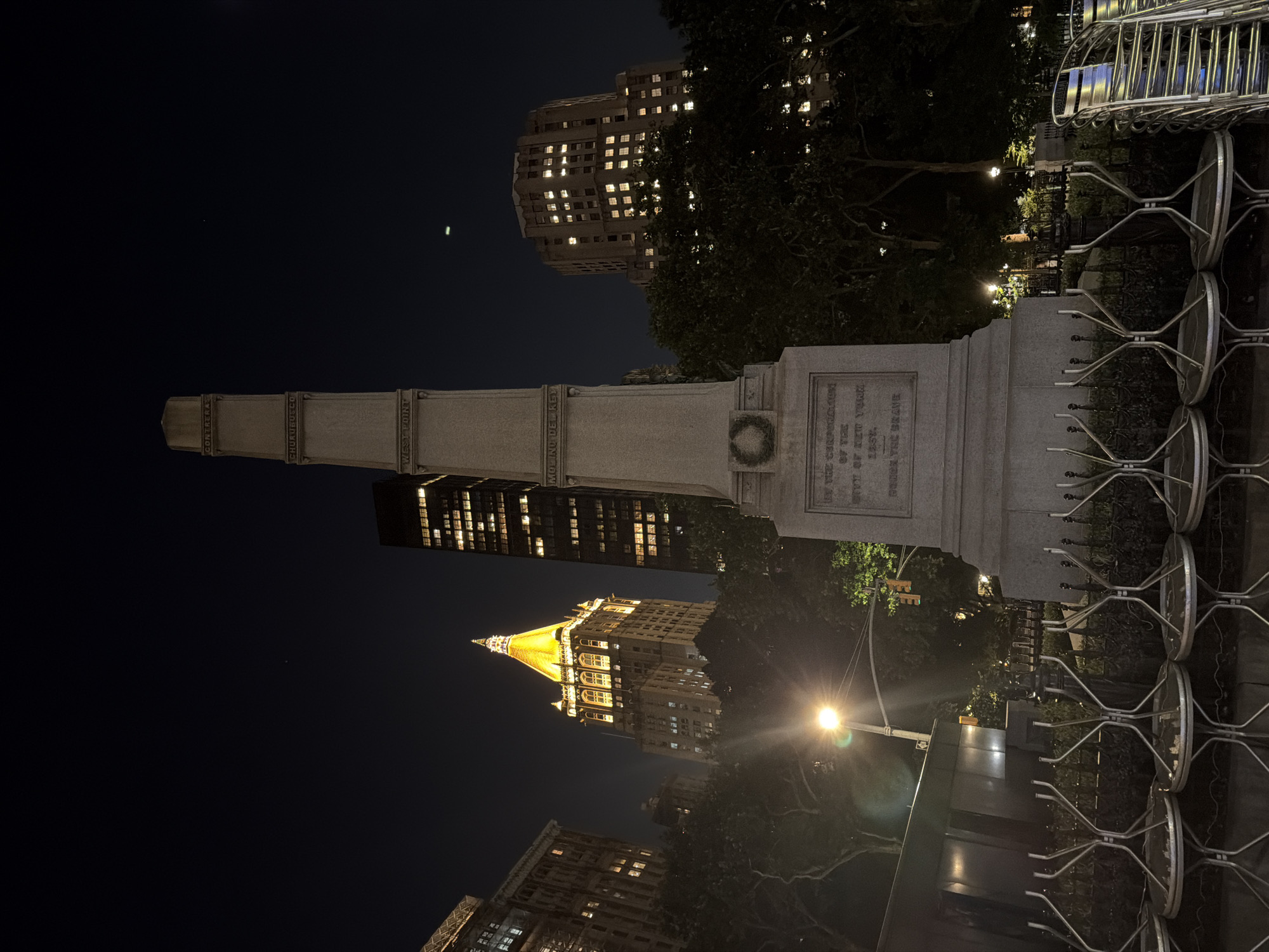 This is a nighttime, low-angle, outdoor shot of a monument in New York City. The monument is made of stone and has inscriptions and a wreath carved into it. Behind the monument are trees, some with streetlights illuminating them, and several buildings, including a tall skyscraper with a bright yellow, illuminated spire, and other buildings with lit windows. In the foreground, there are several tables and chairs stacked up. The sky is dark, with a few faint stars visible. The overall scene appears to be a public square or park in Manhattan.