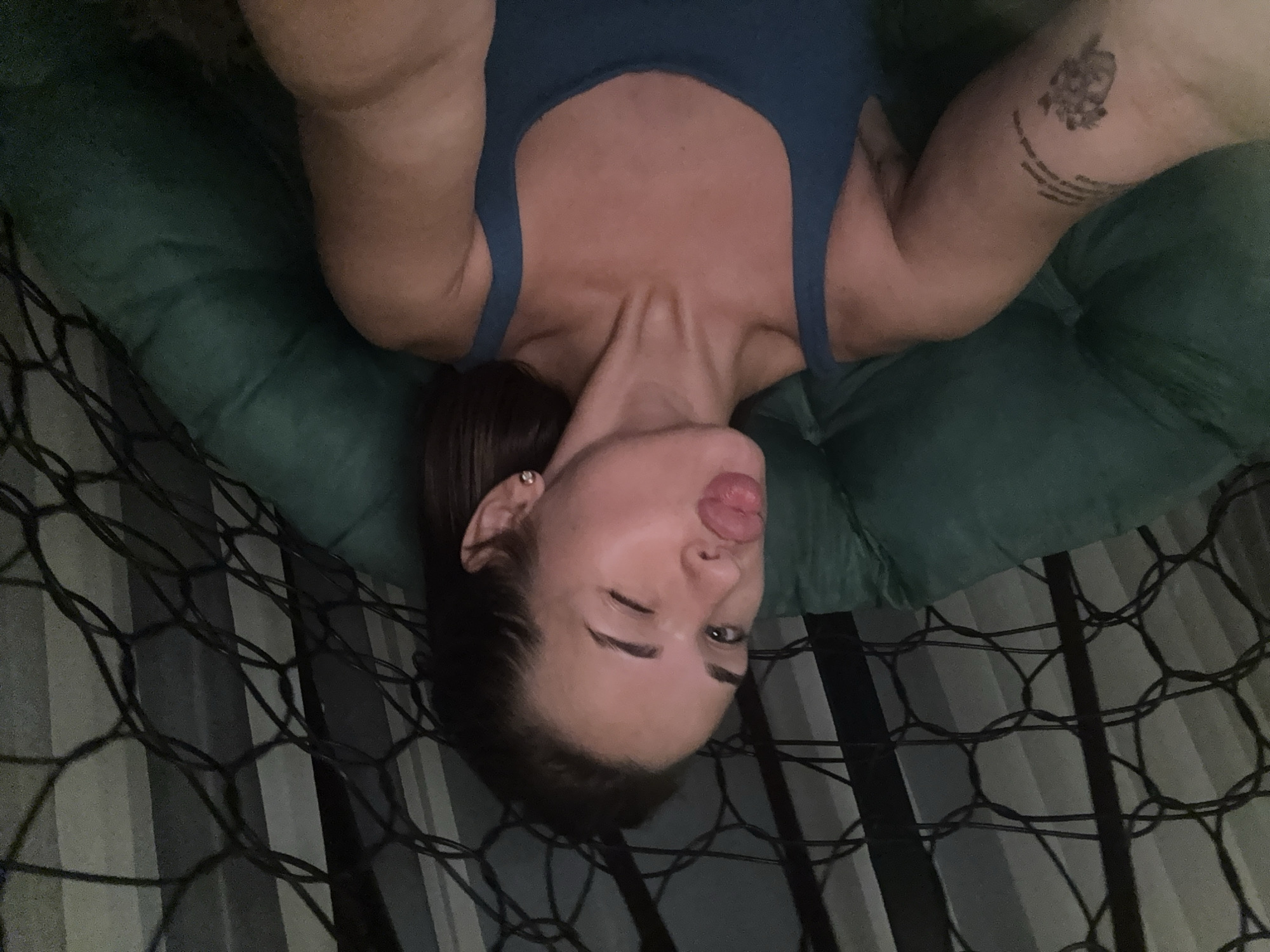 A woman is taking a selfie while sitting in a hanging chair. She is wearing a blue tank top and has a tattoo on her left arm. She is making a kissing face and winking. The background appears to be an indoor setting with vertical blinds or curtains. There is no identifiable information to determine the location in the city of Lobanovo, Russia.
