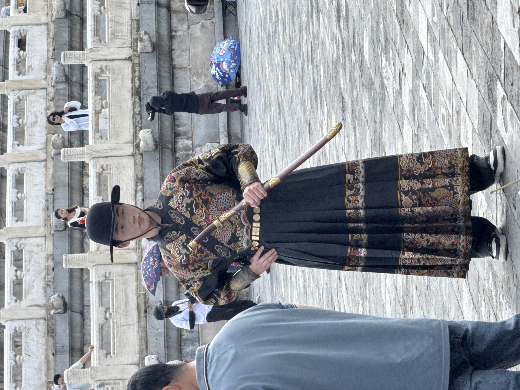 A young East Asian male, appearing to be in his late teens or early twenties, stands in the foreground wearing an elaborate, traditional East Asian costume. The costume is primarily black with intricate gold, red, and blue embroidery depicting mythical creatures, likely dragons. He wears a wide-brimmed black hat and is holding a sword with a gold hilt in his right hand, resting it on the ground. His left hand is on his hip. He is wearing modern white and black sneakers.
In the background, a person in a light blue shirt and jeans is partially visible on the left, facing away from the camera. Behind the central figure, other individuals are present, some holding umbrellas. One person is dressed in white and black and appears to be adjusting their clothing, while another is seen from behind, taking a photograph with a large camera. A blue umbrella with cartoon characters is visible on the ground. The setting appears to be a large, open, paved area in front of ornate, stone structures with multiple levels of tiered seating or walkways. These structures, characterized by white stone balustrades and decorative carvings, suggest a significant historical or cultural site. The pavement is made of large, rectangular grey stones. The overall scene suggests a tourist area, possibly the Forbidden City in Dongcheng District, China, with the central figure possibly part of a historical reenactment or performance. The lighting suggests an overcast day.