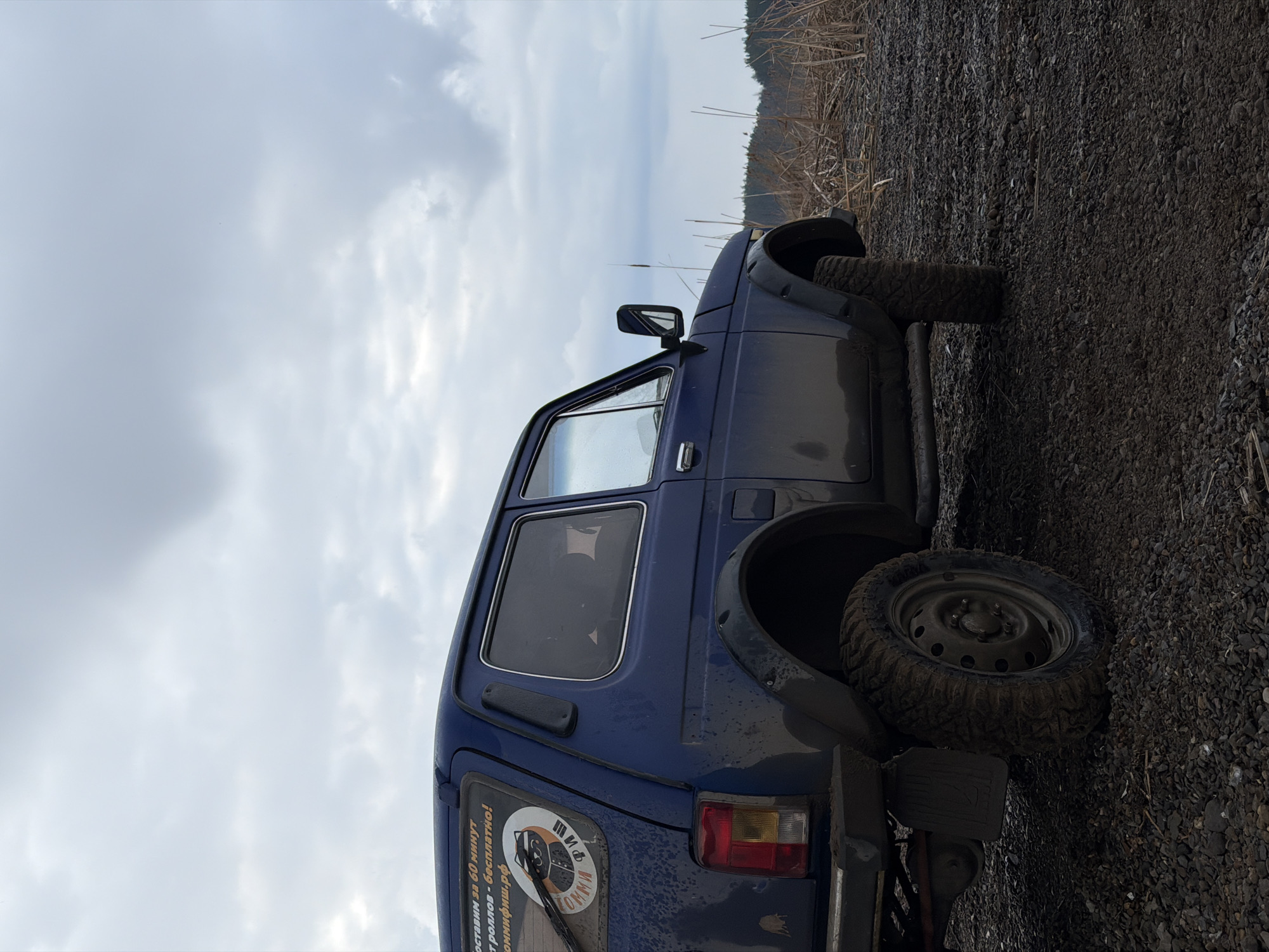 A dark blue, rugged SUV, likely a Lada Niva, is parked on a gravelly and muddy track in an outdoor setting. The vehicle is viewed from its rear-left side, showcasing its dirty exterior with mud splatters on the body and aggressive, knobby, mud-caked tires, indicative of off-road use. A spare tire is mounted on the rear.
The background features a landscape under a heavily overcast sky. Tall, dry reeds or grass are visible along the edge of the track, extending towards a distant treeline or hills that meet the cloudy horizon. The scene suggests a remote or rural environment, possibly near a body of water or marshland. The lighting is dim, consistent with a cloudy day.
A circular sticker on the SUV's rear window displays Cyrillic text and a graphic resembling a stylized fish or animal. The text reads "ПИВ КОМНИ" and "ДОСТАВИМ ЗА 60 МИНУТ БЕСПЛАТНО!" (DELIVER IN 60 MINUTES FOR FREE!). Below this is the website "коммфиши.рф" (commfishi.rf), indicating an advertisement for a delivery service, possibly for food or beverages. The scene is located in Chaikovsky, Russia. No people are visible.