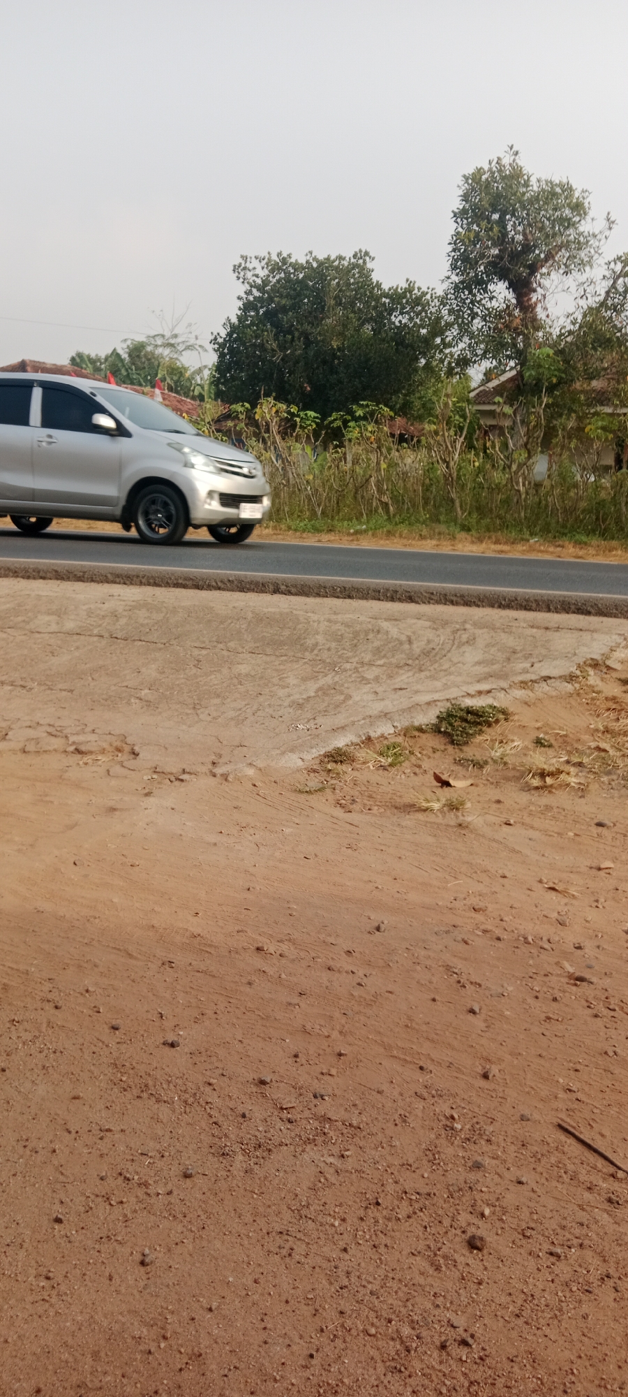 A silver sedan is captured in motion on a paved road in Gondang Rejo, Indonesia. The car is oriented towards the left of the frame, with its front facing partially towards the viewer. Its headlights are on, indicating it is moving. The road surface is a dark asphalt, bordered by a rough concrete embankment on the right. The background features a dense line of trees and shrubbery, with a glimpse of a building with a tiled roof visible among them. The sky is overcast and a pale grey. The foreground consists of dry, reddish-brown soil with scattered pebbles and dried vegetation. The scene suggests a rural or suburban environment on a bright, though cloudy, day.