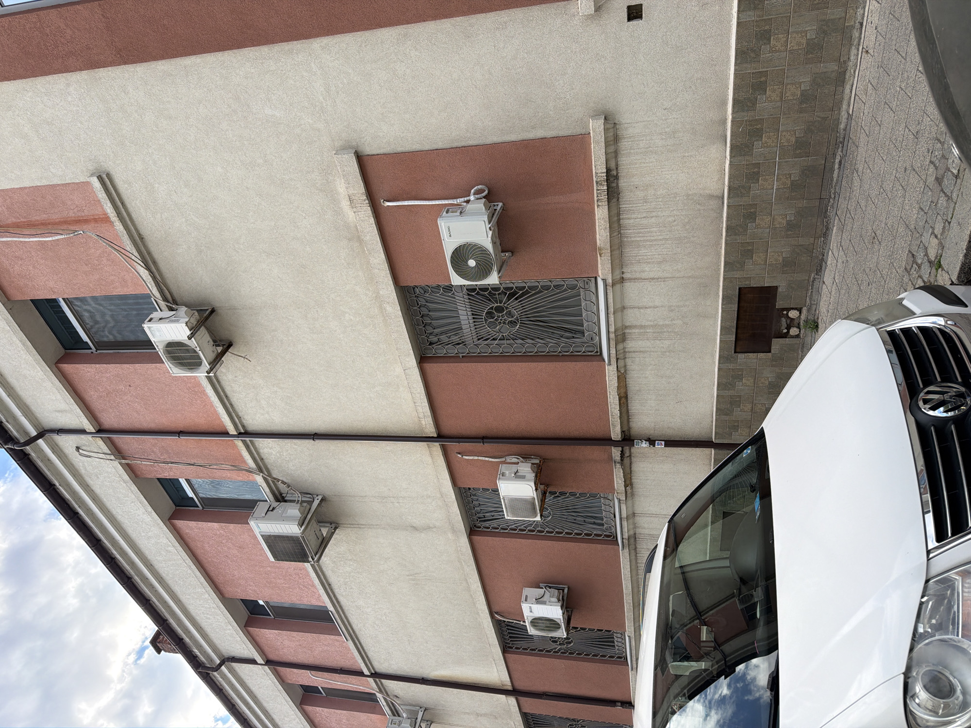 The image shows a white Volkswagen Touran parked on a cobblestone street in front of a building in Plovdiv, Bulgaria. The building is a multi-story structure with a facade of beige and reddish-pink stucco. Several air conditioning units are mounted on the exterior walls of the building. The windows on the lower level are covered with decorative metal grates. The sky above is cloudy.