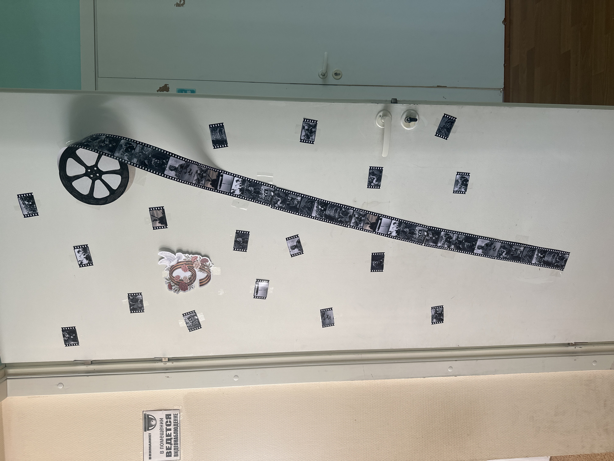 The image shows a white door decorated with a black film reel and a strip of black and white film. There are also several small black and white film frame-like images scattered around the reel and film strip. A sticker depicting a wreath with flowers and a dove is also visible. At the bottom of the door is a warning sign in Russian about video surveillance. The image was taken in Chaikovsky, Russia.