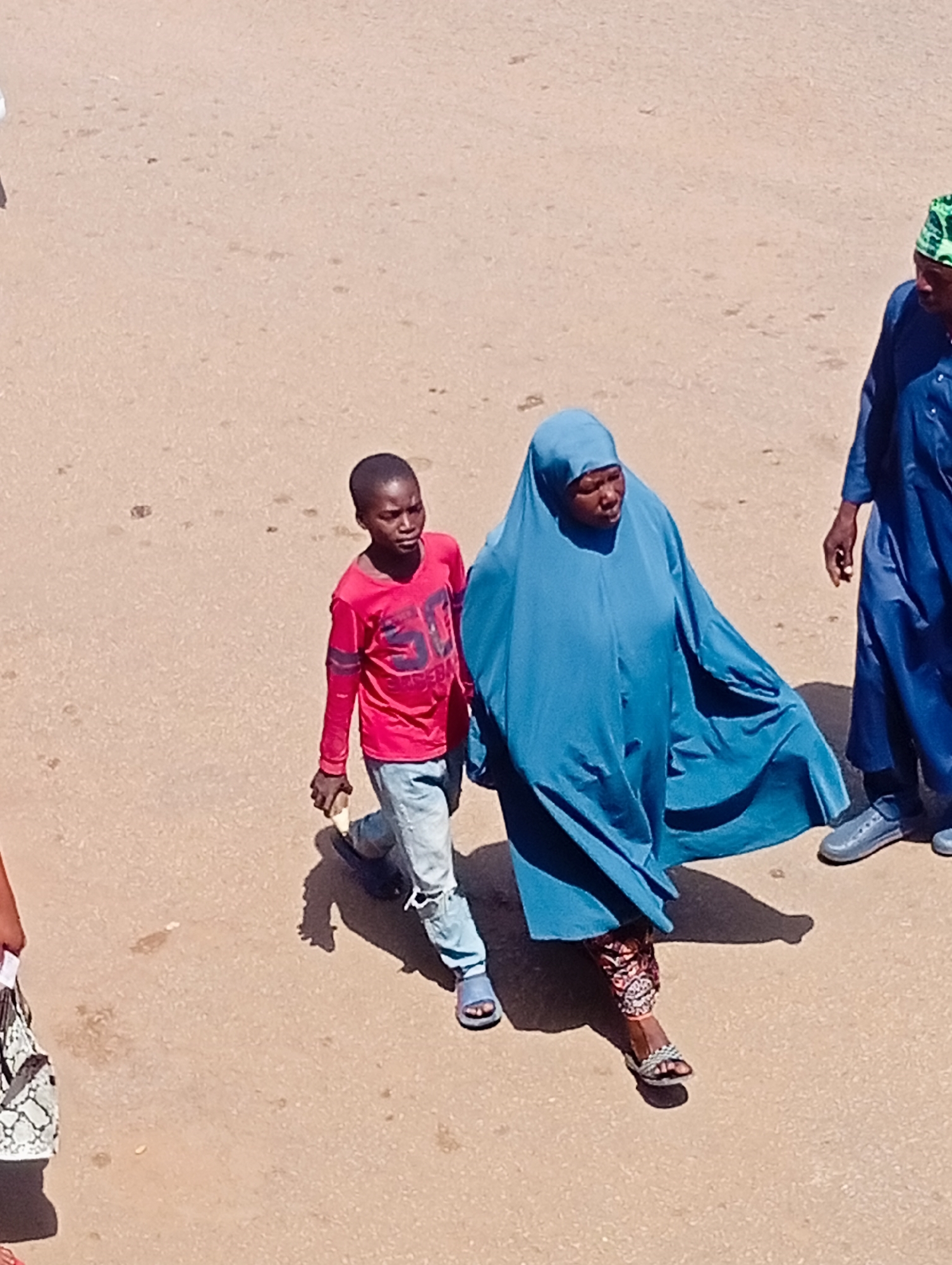 The image captures an aerial view of three people walking on a paved surface, likely a street or courtyard in Bukuru, Nigeria. A young boy in a red long-sleeved shirt and blue jeans walks ahead and to the left. Beside him, and slightly behind, a woman is dressed in a flowing blue hijab that covers her head and body, with colorful patterned pants visible beneath the fabric. To the far right, another person in a blue robe and a patterned green cap is partially visible. The ground appears to be dusty and sandy, suggesting a dry, possibly warm environment. The sunlight casts distinct shadows, indicating it is likely daytime. The overall scene appears to be a casual moment of people walking, perhaps to a market or to a destination. There is no visible text in the image.