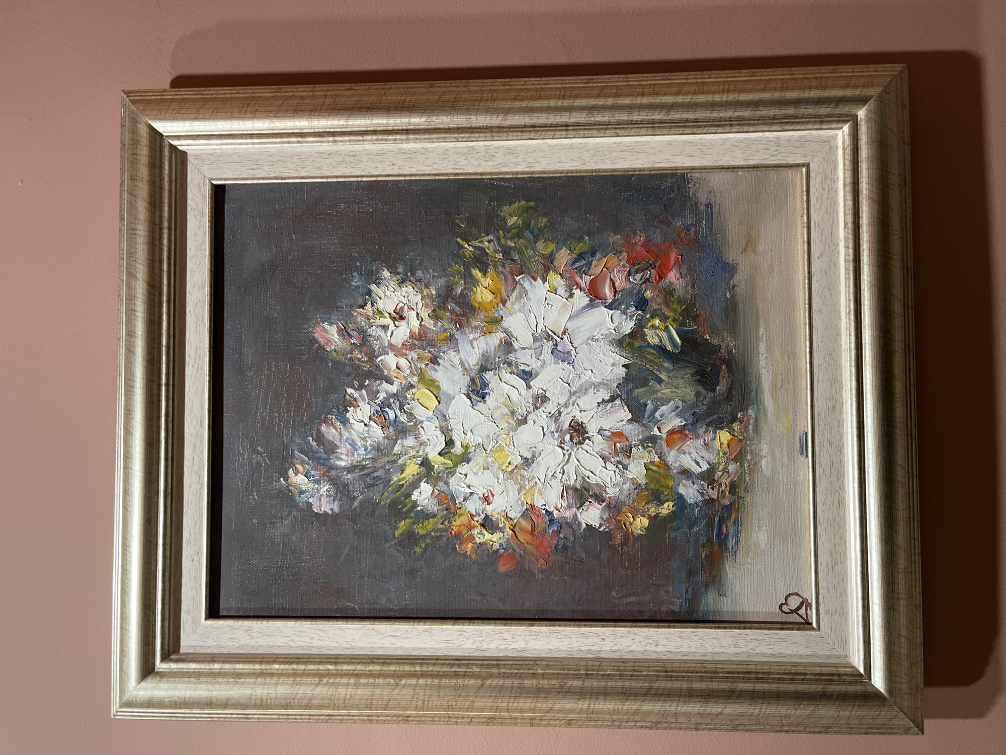 A framed oil painting hangs on a pink-toned wall. The frame is multi-layered and finished in a light, possibly metallic color. The painting itself is of a bouquet of flowers rendered in thick impasto strokes, predominately white with touches of yellow, red, green, and blue against a dark, gray-brown background. A small, heart-shaped signature is visible in the lower right corner of the painting. No people are present in the image. The location of Plovdiv, Bulgaria is not present in the image.