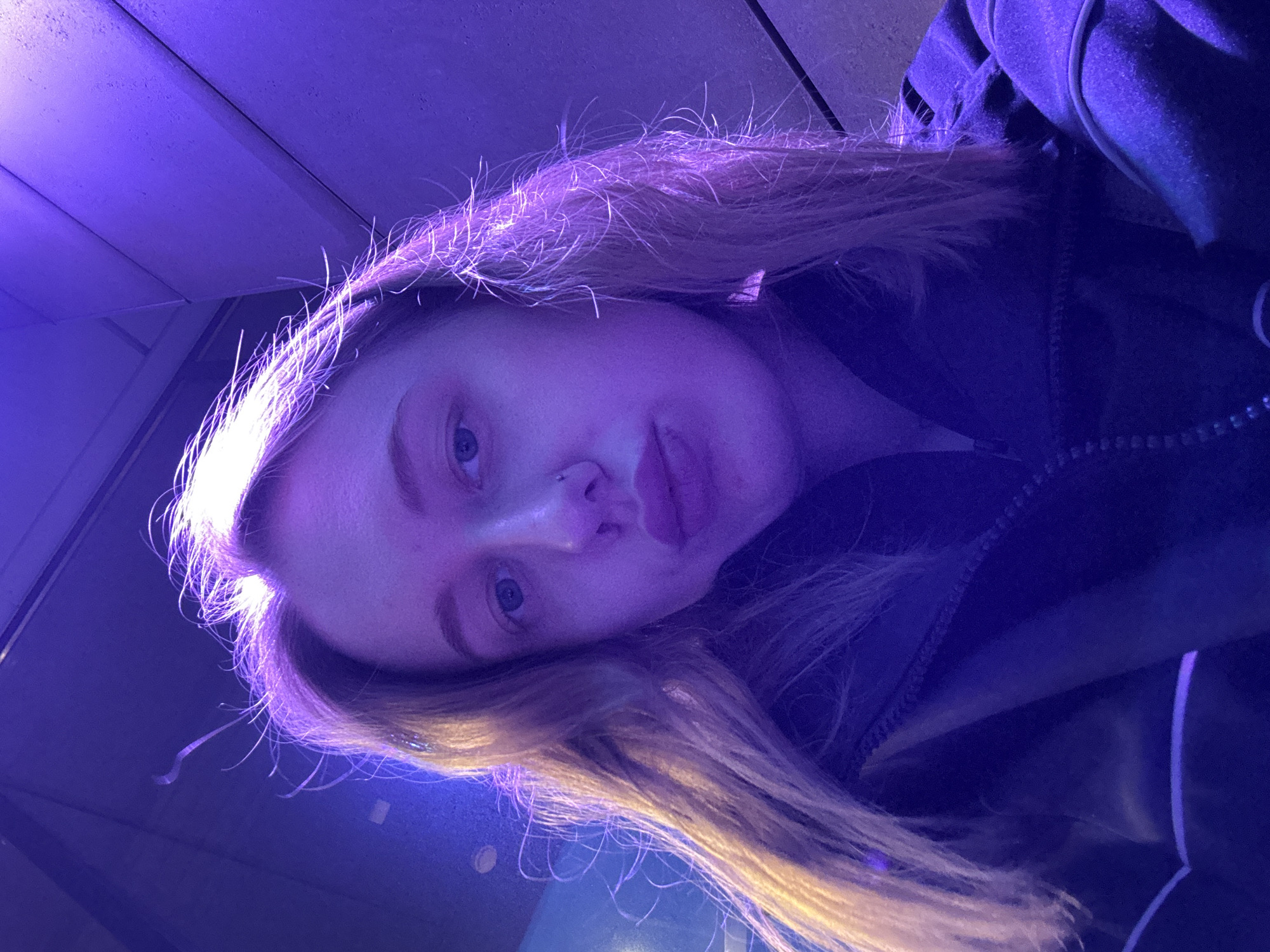 A close-up, portrait-style photograph captures a young woman with fair skin and light brown or blonde hair, illuminated by purple and blue lighting. Her face is slightly tilted, with her eyes open and directed towards the camera. She appears to be wearing a dark jacket. The background is blurred, showing a ceiling with paneling. No specific details indicating the location of Ol'ginskaya, Russia are visible. The image focuses on the woman's face and immediate surroundings.