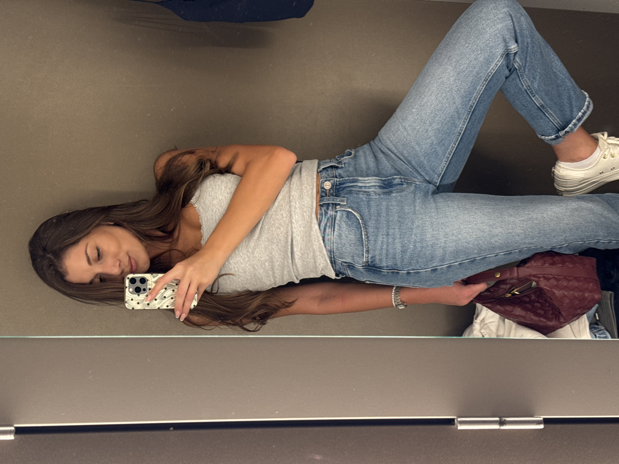 A young woman lies on her back in a dressing room, taking a selfie with her smartphone. She is wearing a gray tank top and light wash jeans, with white sneakers. A burgundy woven handbag is near her right leg. The room is a dressing room. The image doesn't contain any information to relate to the city of Burgas, Bulgaria.