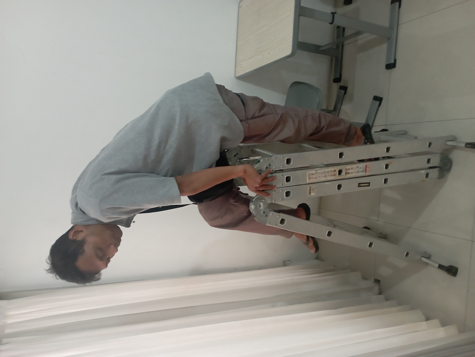 A man is on a ladder in a room with white walls and white curtains. He is wearing a grey t-shirt and brown pants. The ladder is silver and appears to be an extension ladder. There is a small desk with a light wood top and grey metal legs in the background. A grey chair is partially visible. The floor is tiled with a light grey color. The scene is likely indoors.