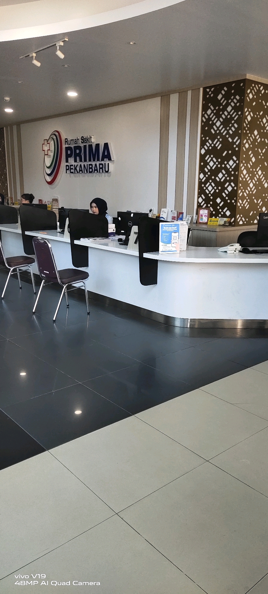 The image shows the interior of a hospital reception area in Pekanbaru, Indonesia. A white reception desk stretches across the frame, with two female employees seated behind it, facing computer monitors. One employee, wearing a black hijab, is actively engaged with her computer. Several chairs are positioned in front of the desk, indicating a waiting area. The hospital's logo, which reads "Rumah Sakit PRIMA PEKANBARU," is prominently displayed on the wall behind the receptionists. The floor is tiled with a contrasting pattern of dark gray and light beige squares. The overall environment appears clean and modern, suggesting a functional and professional setting.