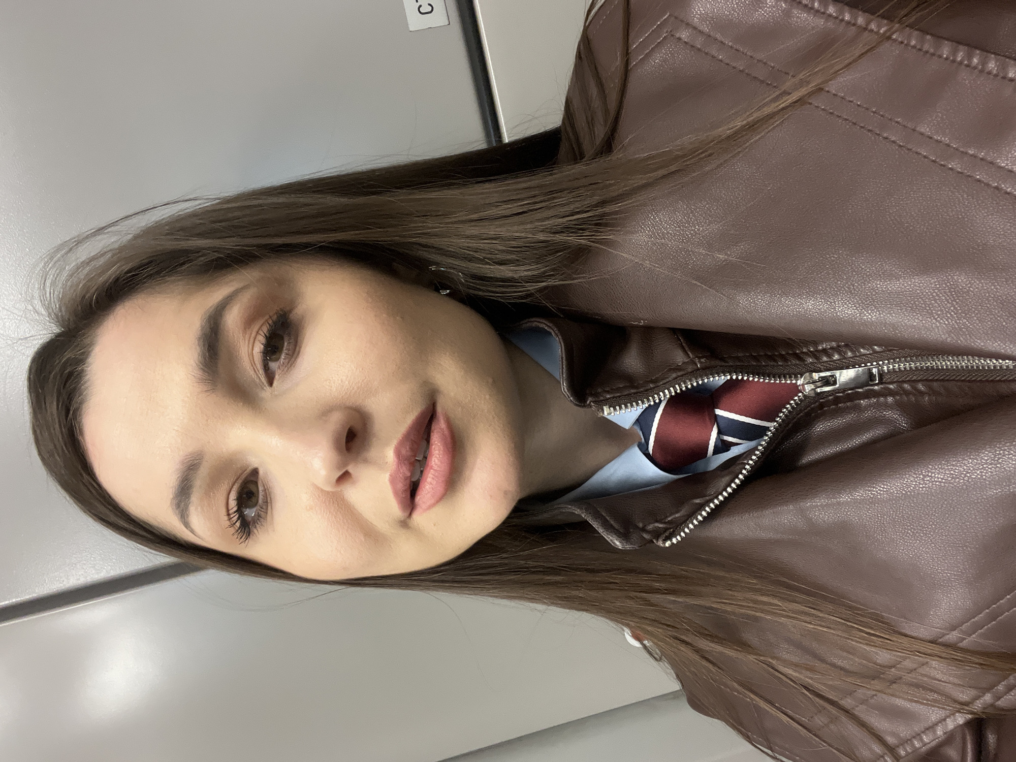 A close-up portrait shows a young woman with long, dark brown hair, looking directly at the camera. She is wearing a brown leather jacket, which is partially unzipped. Beneath the jacket, a light blue shirt is visible, along with a tie featuring diagonal stripes in dark red, navy, and white/light blue. Her face displays subtle makeup, including mascara on her eyelashes, defined eyebrows, and a light pink lip color; her lips are slightly parted, revealing her teeth. The background consists of plain grey panels, suggesting an indoor setting. A white label with a black letter "C" is visible in the upper left background. The woman's expression is calm and neutral. The location context provided is Moscow, Russia.