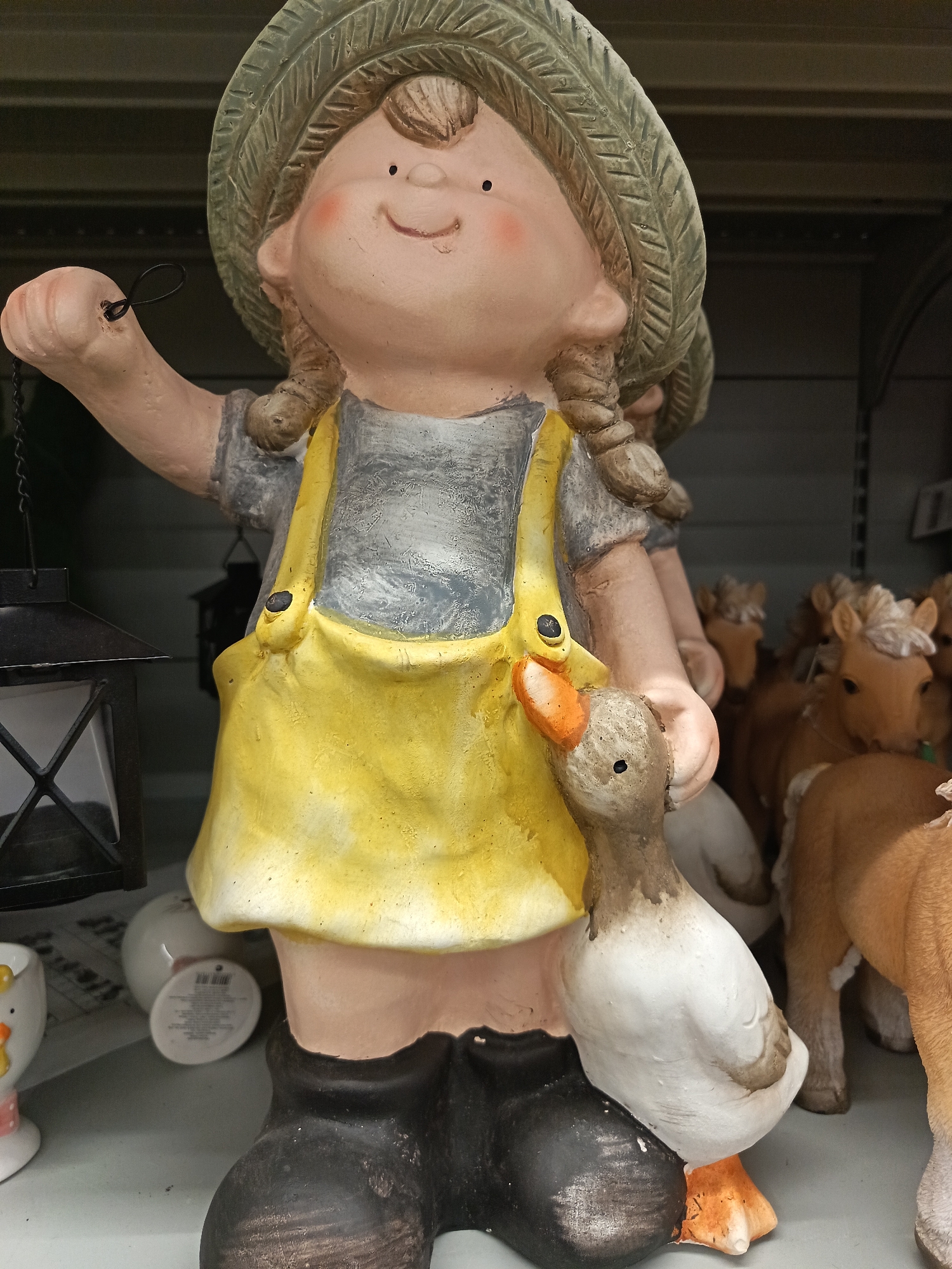 A ceramic figurine of a girl in a straw hat and yellow overalls is standing on a shelf, holding a lantern in her left hand and a duck in her right. The girl has rosy cheeks and braided pigtails. Behind her are figurines of toy horses and other ceramic objects. The location is likely a store or a display area and not necessarily located in the city of Nizhnevartovsk, Russia.