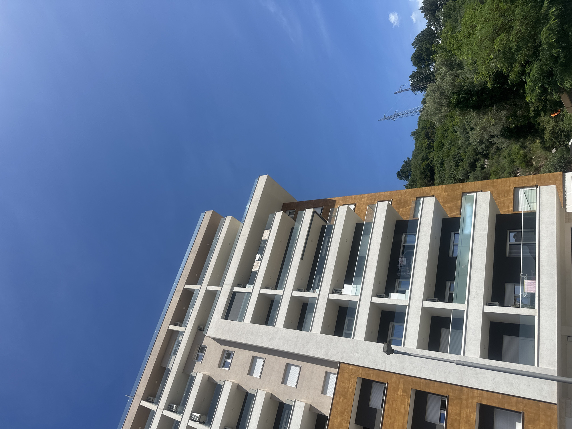 This image showcases a modern, multi-story building against a clear blue sky, flanked by a lush green hillside on the right. The building exhibits contemporary architecture with a facade composed of light beige or off-white stucco alternating with prominent sections of rich, dark brown, possibly wood or metal panelling. Numerous recessed balconies line the structure, each featuring transparent glass railings. Several outdoor air conditioning units are visible within these balcony spaces, and on one balcony, clothes are hanging on a drying rack, suggesting residential use.
To the right of the building, a densely forested hillside rises, covered with various green trees. Partially obscured by the foliage, a large metal electricity transmission pylon stands on the hillside, indicating nearby infrastructure. A small, bright orange object is also visible among the trees on the far right. The sky above is a vibrant, nearly cloudless blue, with only a few faint white wisps. The scene is brightly lit by the sun, suggesting daytime, likely morning or early afternoon. A grey streetlight pole extends horizontally across a lower section of the building's facade. The image presents a blend of modern urban development with a natural, green environment, consistent with locations like Budva, Montenegro. No discernible text is visible.