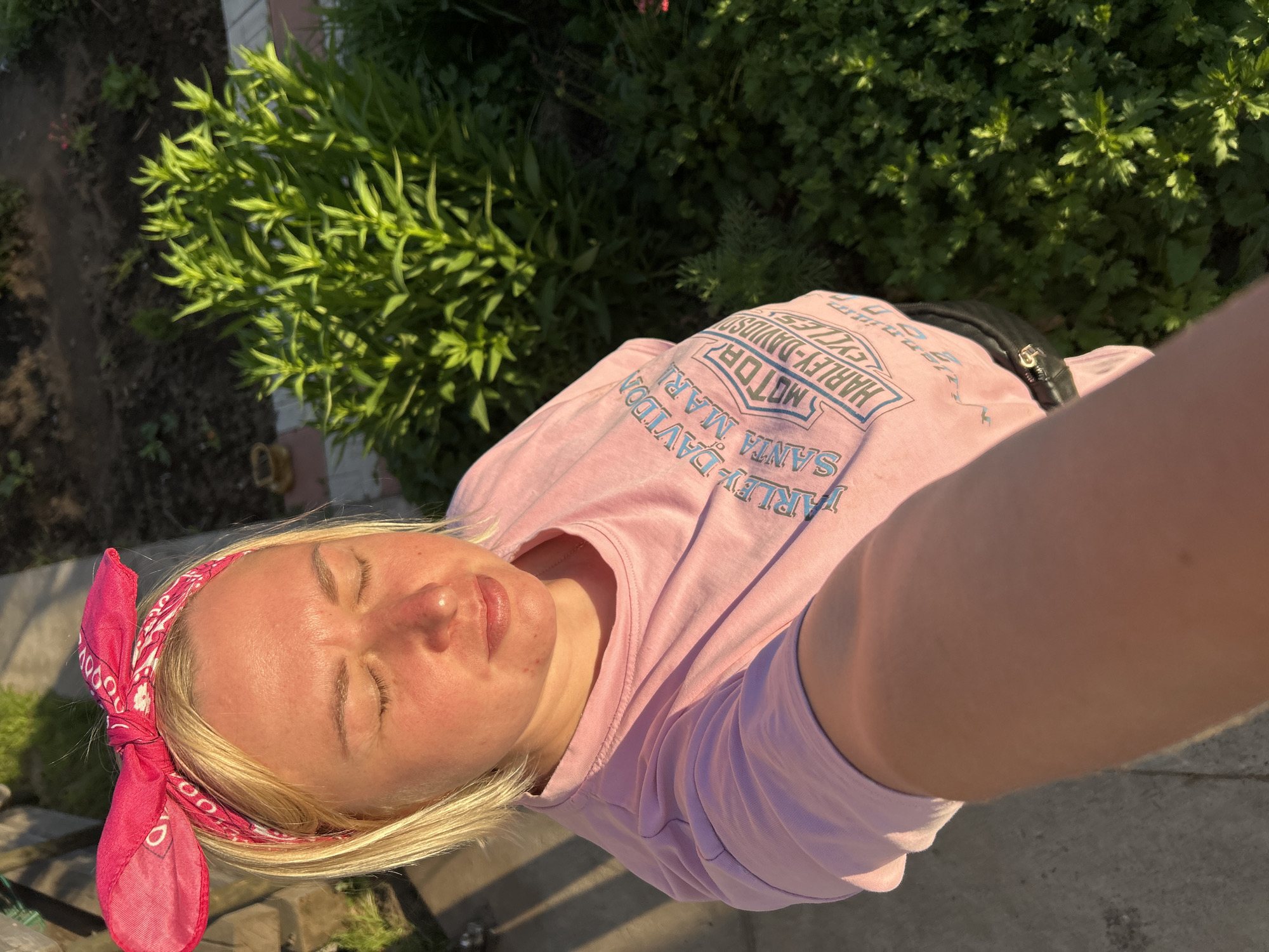A blonde woman with her eyes closed and a pink bandana tied around her head takes a selfie outdoors. She is wearing a pink t-shirt with "HARLEY-DAVIDSON SANTA MARIA CYCLES" written across it in blue lettering. The background shows a garden with green plants and a paved path. The sunlight casts shadows on the ground. This scene is consistent with a residential area in Chaikovsky, Russia, possibly a private garden or backyard.