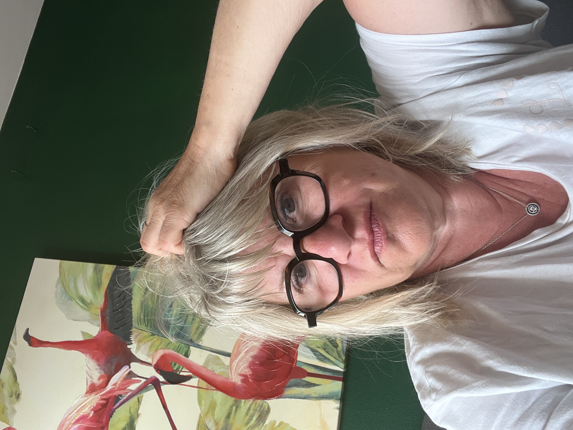 A woman with fair skin and blond hair is looking at the camera. She is wearing black-framed glasses, a white t-shirt, and a silver necklace. Her right hand is in her hair. Behind her is a green wall and a painting of pink flamingos. The location cannot be determined from the image.