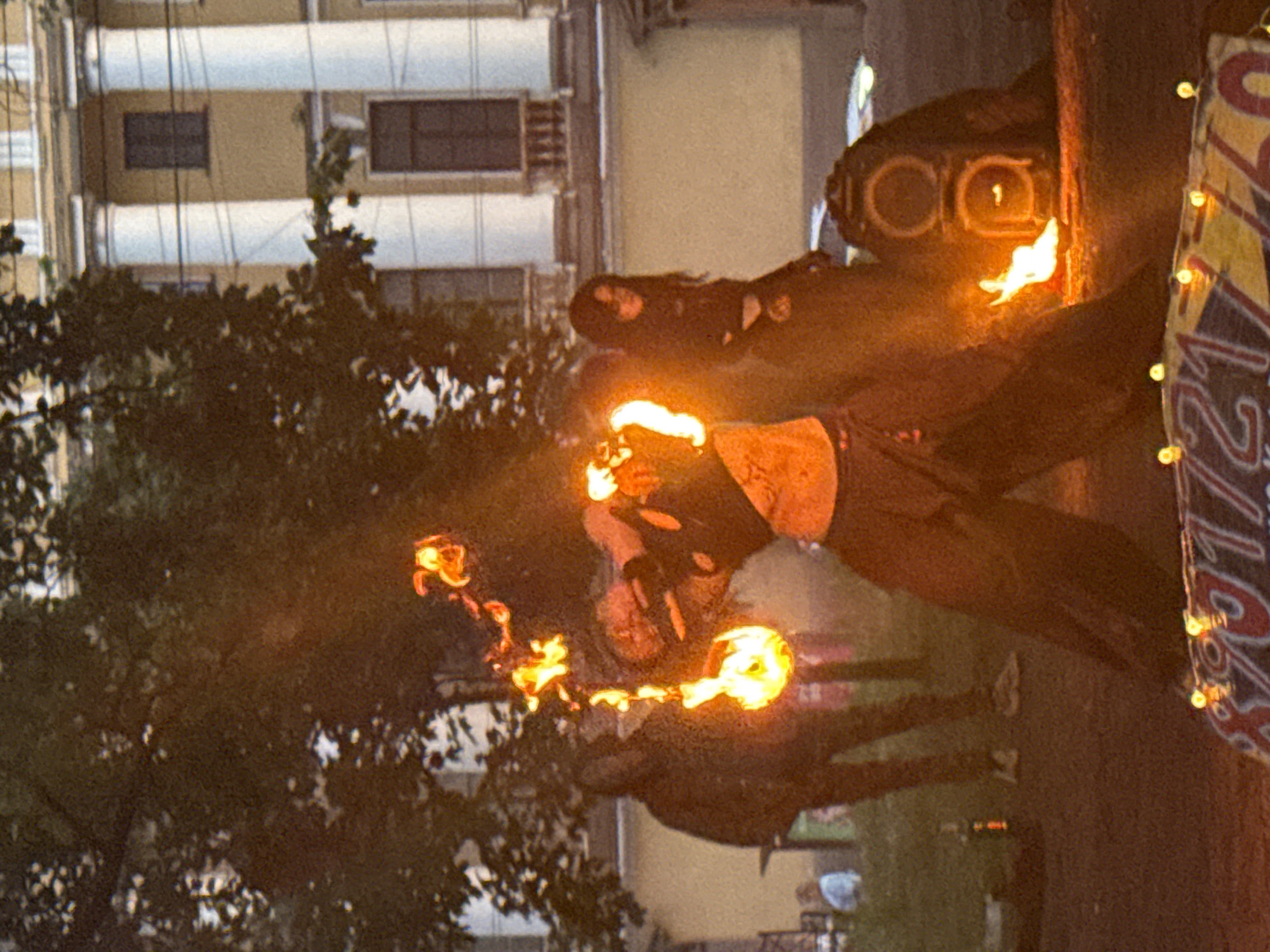 The image captures a street performance in front of a building with classical architecture, likely in Saint Petersburg, Russia, given the style of the building which features columns and arched windows. In the foreground, a performer is engaged in fire spinning, holding a flaming object that trails fire. The performer is dressed in dark clothing, with visible fire effects around their hands and body. To the right of the main performer, another individual, possibly a woman with dark hair, stands observing the performance. In the background, the facade of a building with multiple windows and a distinctly European style is visible, illuminated by natural light. The ground appears to be paved or dirt, with some scattered objects. A speaker system is partially visible to the right of the performer, suggesting the event might involve music or amplification. The overall atmosphere suggests a public outdoor event, possibly a festival or a spontaneous street performance.