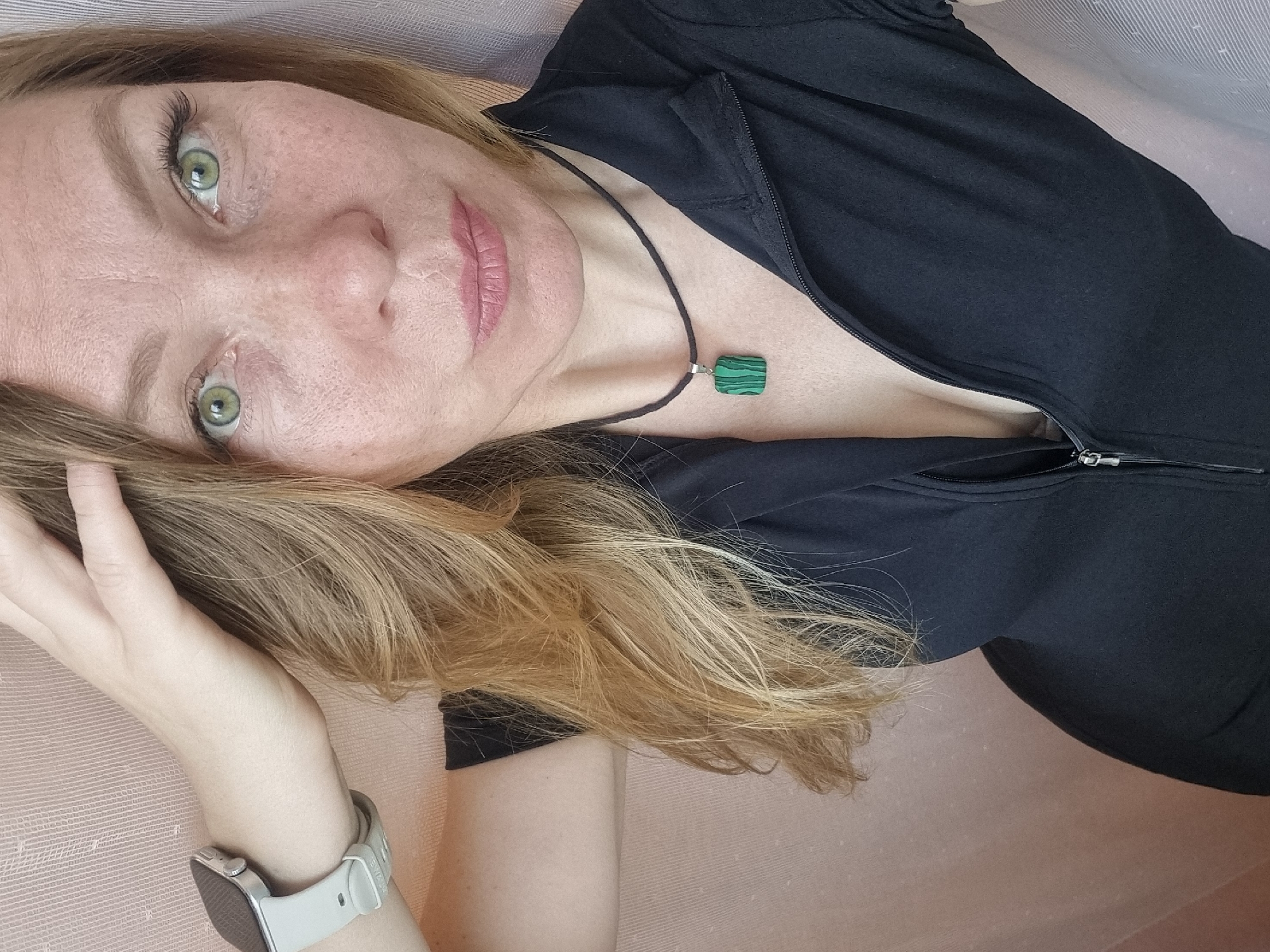 A woman with light brown hair and green eyes is lying down, looking up at the camera. She is wearing a black top with a zipper and a green square pendant necklace. She has freckles on her face and is wearing a light-colored smartwatch on her left wrist. Her left hand is near her face, touching her hair. The background appears to be a light pink fabric. There is no indication in the image that it was taken in Chaikovsky, Russia.