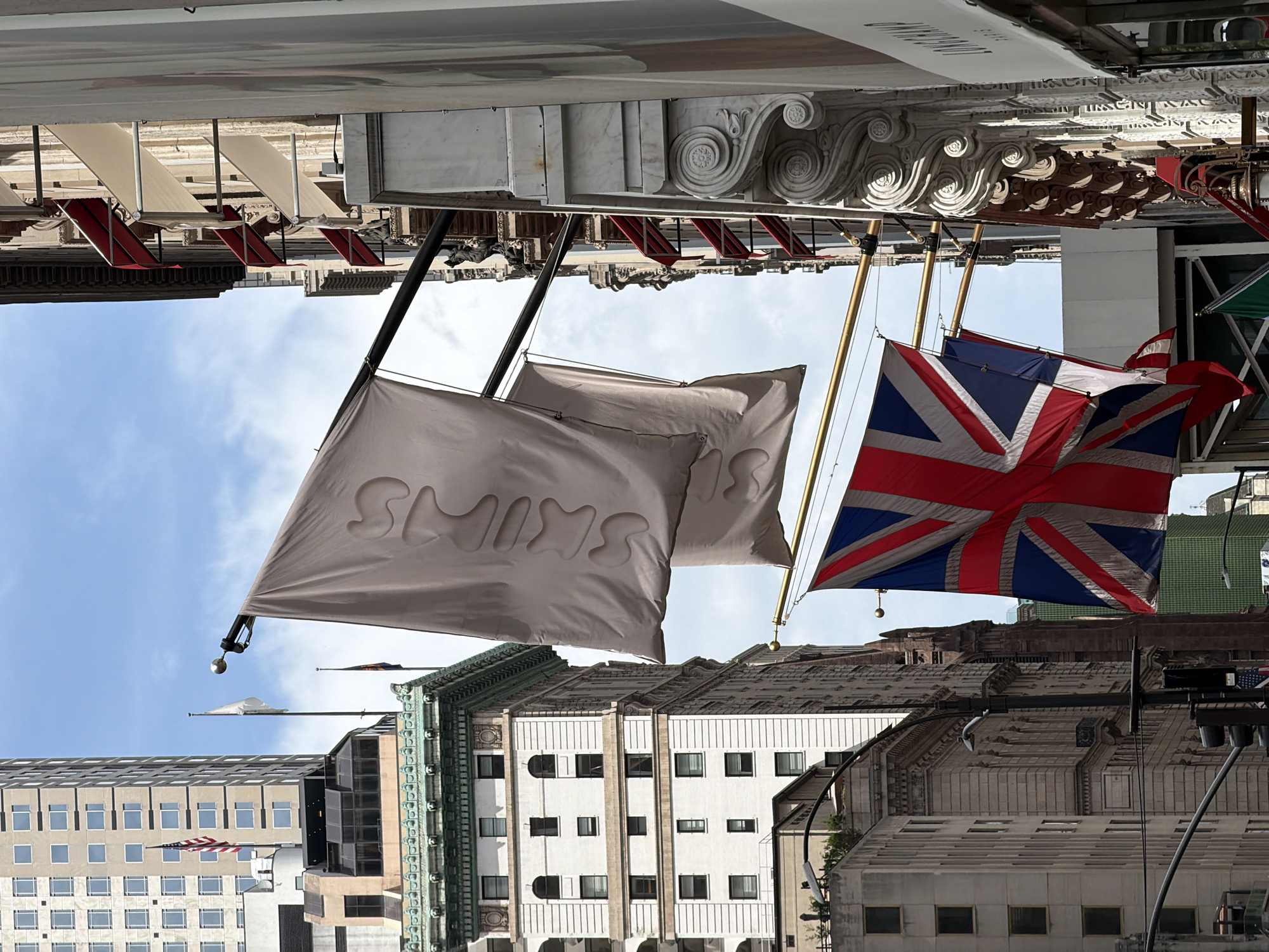 This is a photograph taken on Fifth Avenue in New York City, with the Empire State Building visible in the background. The image captures a street scene dominated by tall buildings and flags.
In the foreground, a large, light-colored flag with embossed text is displayed, partially obscuring the view of a tall, modern building on the left. Below this, the Union Jack flag is prominently featured, flying from a pole. Further down, glimpses of other flags are visible. The buildings on the right are ornate, with decorative stonework and red and cream awnings. The sky is partly cloudy. The overall impression is one of an urban environment with a touch of grandeur.