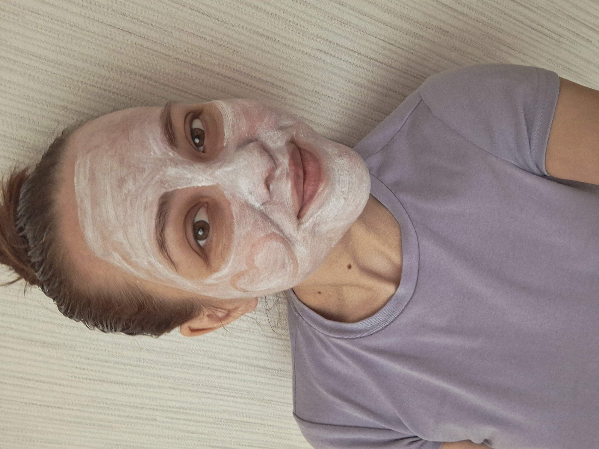 A woman with a white facial mask is lying down, facing the camera. Her hair is pulled back, and she is wearing a light purple t-shirt. She is lying on a textured surface. The location is the city of Chaikovsky, Russia.