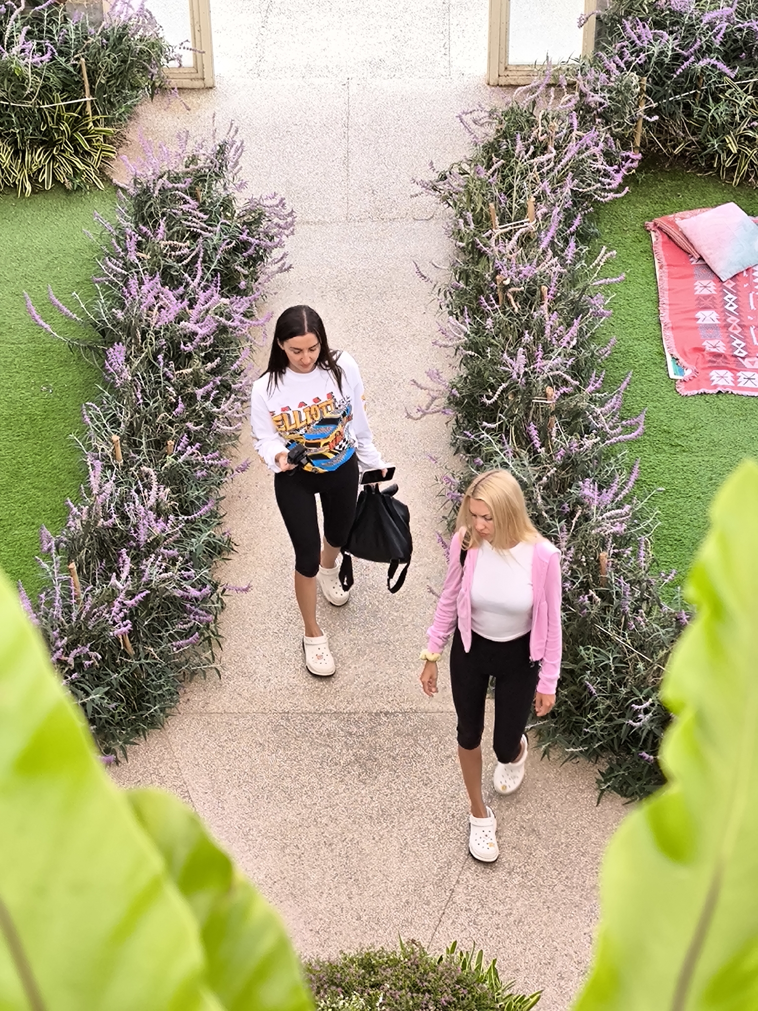 Two women walk along a stone pathway lined with lavender plants and green turf. The woman closest to the foreground wears a pink cardigan over a white top and black leggings, with white clogs on her feet. The other woman wears a white long-sleeved shirt, black leggings, and white clogs. She also carries a black bag. A picnic blanket and pillow sit on the green turf next to the pathway, where the background includes a beige wall with a window. A green leaf obscures the foreground view from the bottom. The setting appears to be an outdoor area in Candikuning, Indonesia.