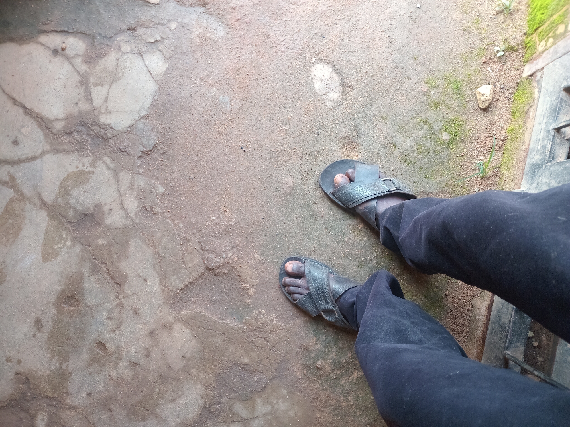 The image shows a pair of black sandals on a person's feet, wearing dark pants. The sandals are made of leather with a criss-cross strap design. The ground beneath is a rough, cracked, and somewhat uneven surface, appearing to be made of concrete or packed earth, with patches of dry soil and sparse green moss. The scene is lit from above, casting subtle shadows. The overall environment suggests an outdoor or semi-outdoor setting, possibly a pathway or yard. The provided crops offer close-ups of the ground texture and the person's feet and attire, highlighting details like the cracked surface and the material of the sandals. The location is identified as Bukuru, Nigeria.