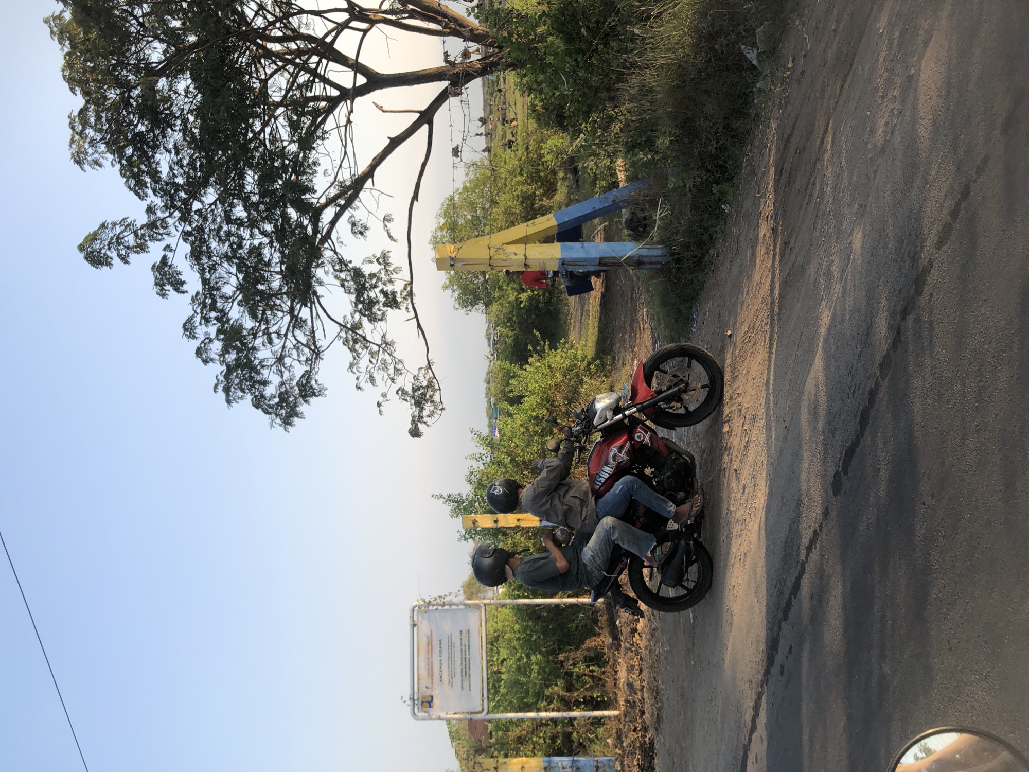 Two individuals wearing helmets are riding a red motorcycle on a dirt and asphalt road in Gempolsari, Indonesia. The person in the back is driving, and the person in the front is a passenger. They are wearing casual clothing, including jeans and shirts. The motorcycle has a license plate that is partially visible.
In the background, there is a sign with text in Indonesian, which appears to be an official notice. There are also yellow and blue painted posts, indicating some form of barrier or entrance. Lush green vegetation and trees surround the area, with a large tree with dense foliage dominating the right side of the frame. The sky is clear and blue, suggesting it is daytime. The scene is likely a rural or semi-rural area within Gempolsari.