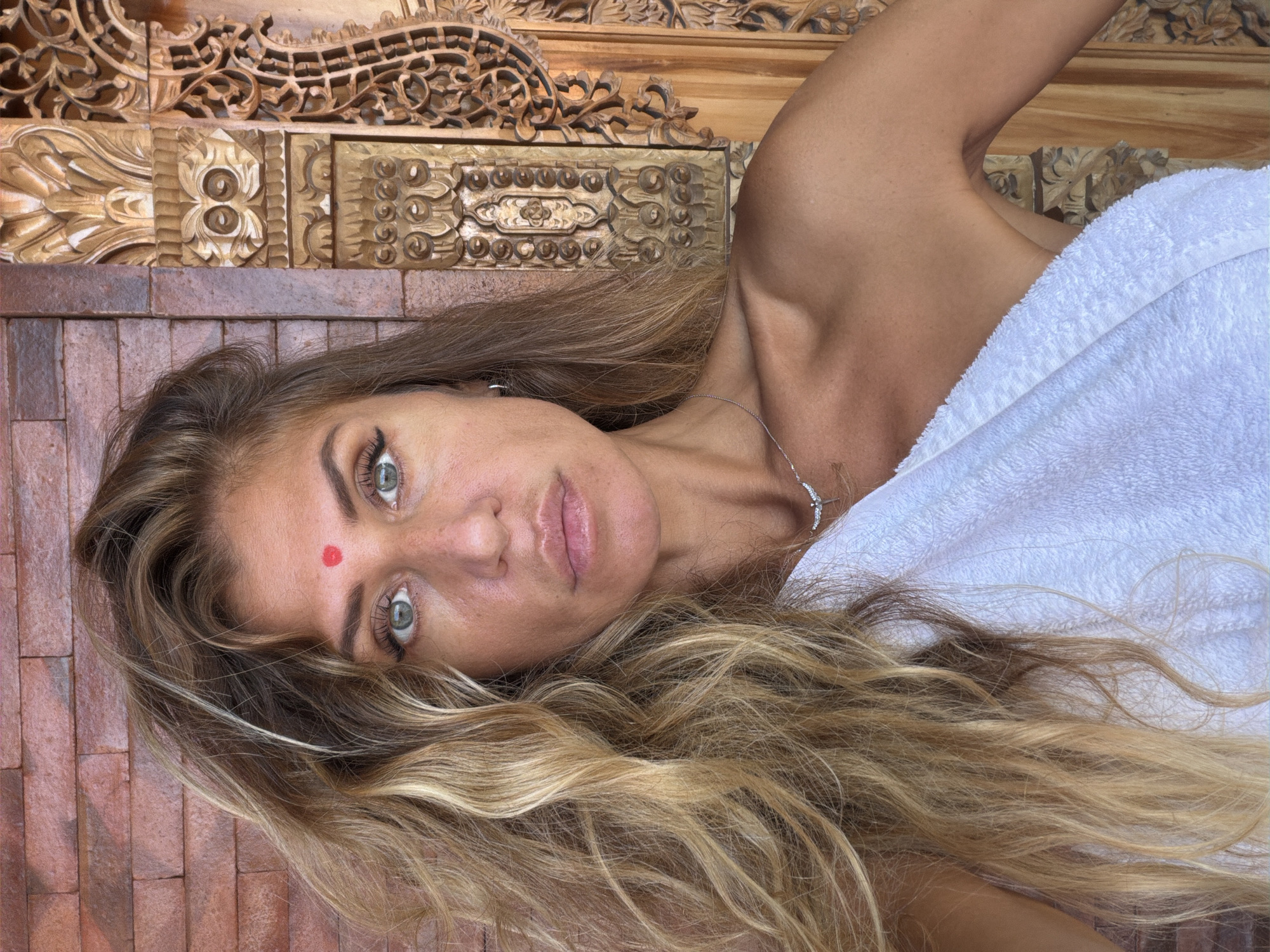 A fair-skinned woman with long, wavy blonde hair and a red bindi on her forehead is pictured from the chest up. She is wearing a white towel draped over her shoulders and a silver necklace. Her eyes are green, and she has dark eyeliner on her upper lids. The background is a textured brick wall on the left and a carved wooden panel on the right. The setting appears to be outdoors, possibly in a tropical location like North Kuta, Indonesia, given the style of the wood carving and the brickwork.