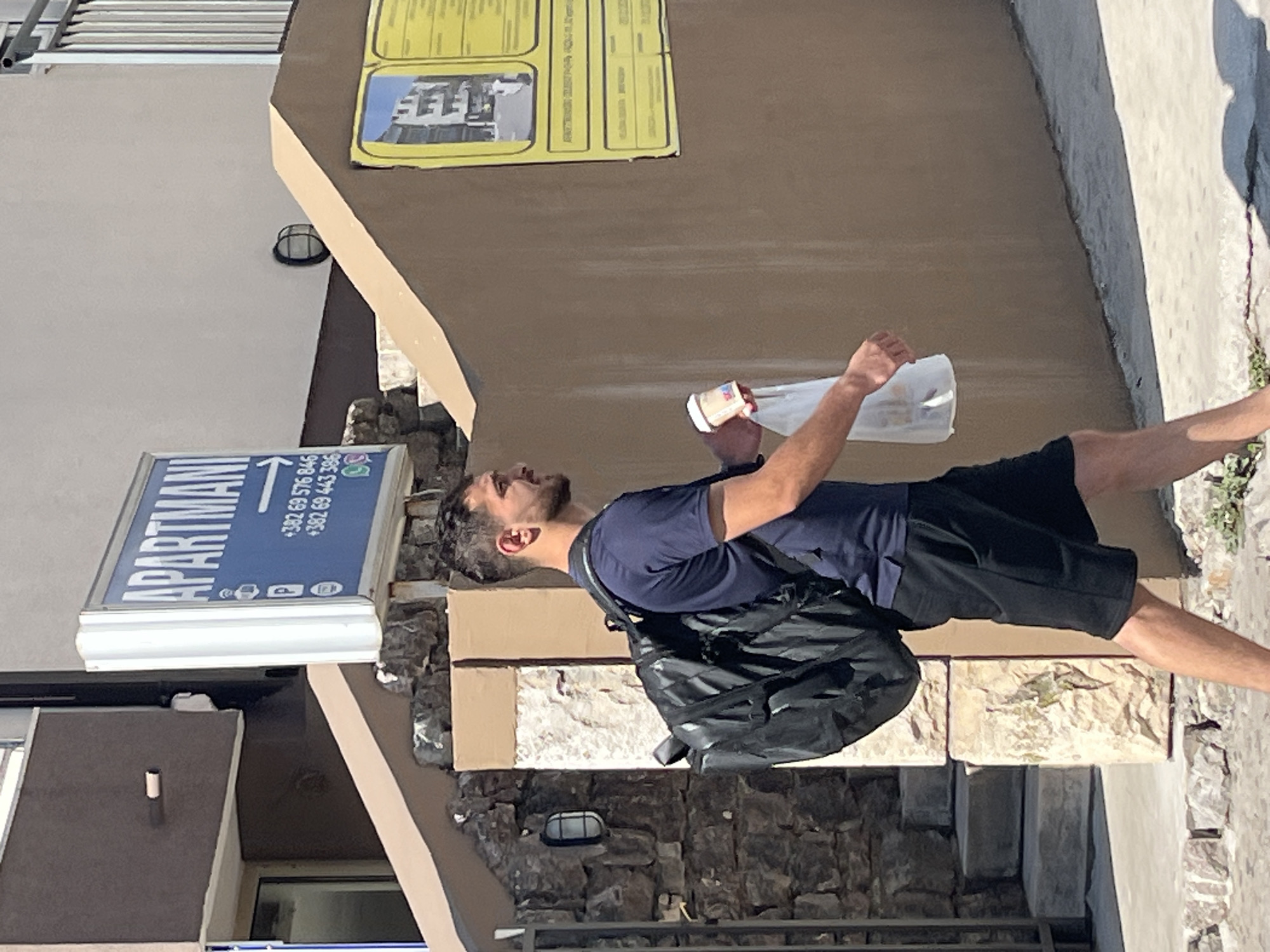 A young man with dark hair and a beard walks from left to right in front of a building with a brown stucco wall. He wears a dark blue polo shirt and black shorts. A large black backpack is on his shoulders, and he holds a white paper cup in his right hand and a white plastic bag in his left. A blue sign that reads "APARTMANI" with phone numbers is visible on the left side of the image, indicating the location is an apartment building. The man appears to be walking on a paved sidewalk. The scene is outdoors in daylight.