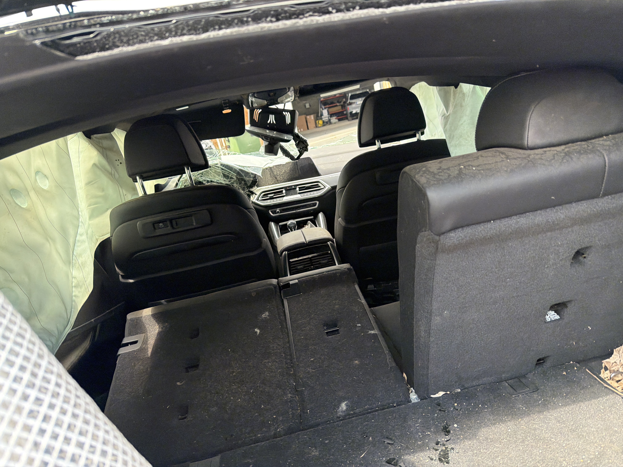 The image shows the interior of a damaged vehicle, likely after a collision. The airbags have been deployed. The windshield is cracked. The interior appears dirty and disheveled. The location cannot be determined from the image. The image contains no people.