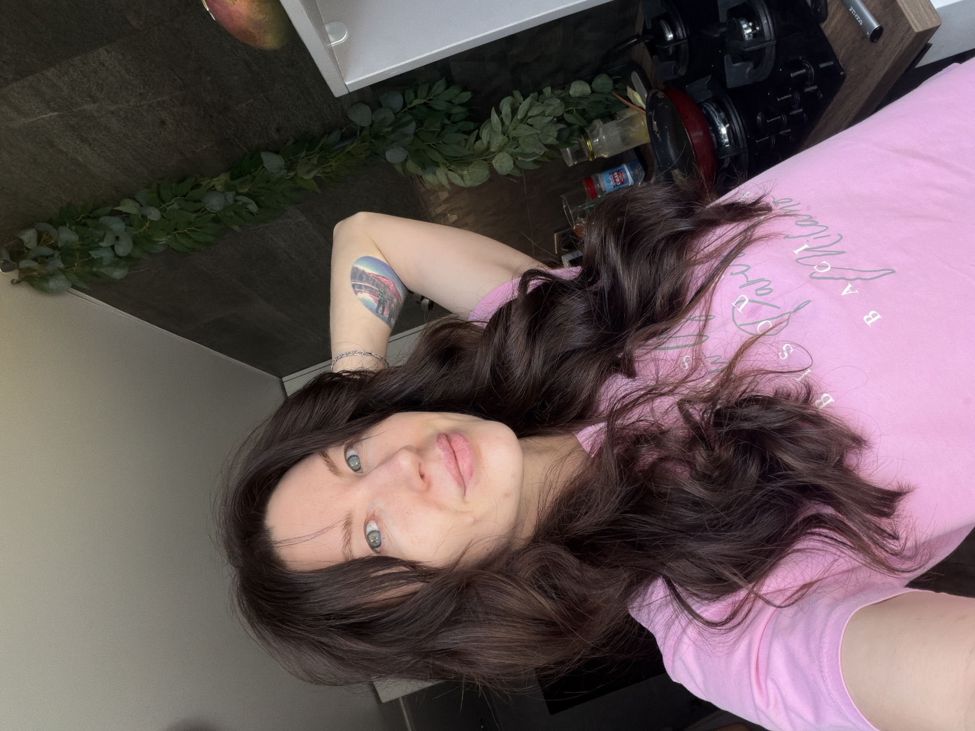 A female individual is depicted in a self-portrait, facing the camera with a slight smile. She has long, dark brown wavy hair, light blue eyes, and light skin, with a small mark visible on her right cheek. She is wearing a pink t-shirt with grey text that partially reads "BACI Paris Milano." Her left arm is raised, with the hand positioned behind her head, revealing a multicolored tattoo on her inner left bicep and a silver chain bracelet on her left wrist.
The setting appears to be an indoor kitchen or kitchenette area. In the background, to the right of the subject, a dark, textured wall features a vertical garland of artificial green leaves. Below this, on a wooden countertop, a black four-burner gas cooktop is visible, with a red frying pan resting on one of the burners. Adjacent to the cooktop are a blue container labeled "СОЛЬ" (SALT) in Cyrillic and a clear jar containing a yellow liquid. A white cabinet or shelf is positioned to the far right, displaying a red and yellow fruit, likely a mango. The ceiling above is light grey.
The specific location within the city of Perm, Russia, cannot be determined from the visual information provided in the image.
